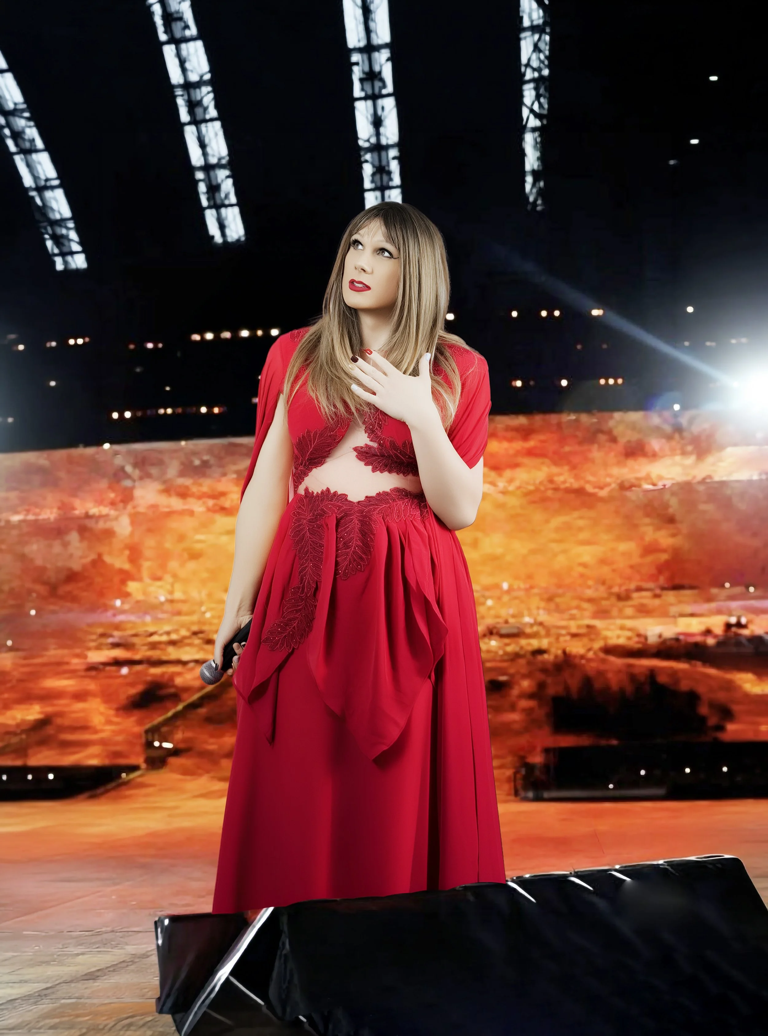 A woman dressed in a red gown with lace details, holding a microphone, stands on a stage with a fiery background and a dark overhead structure.