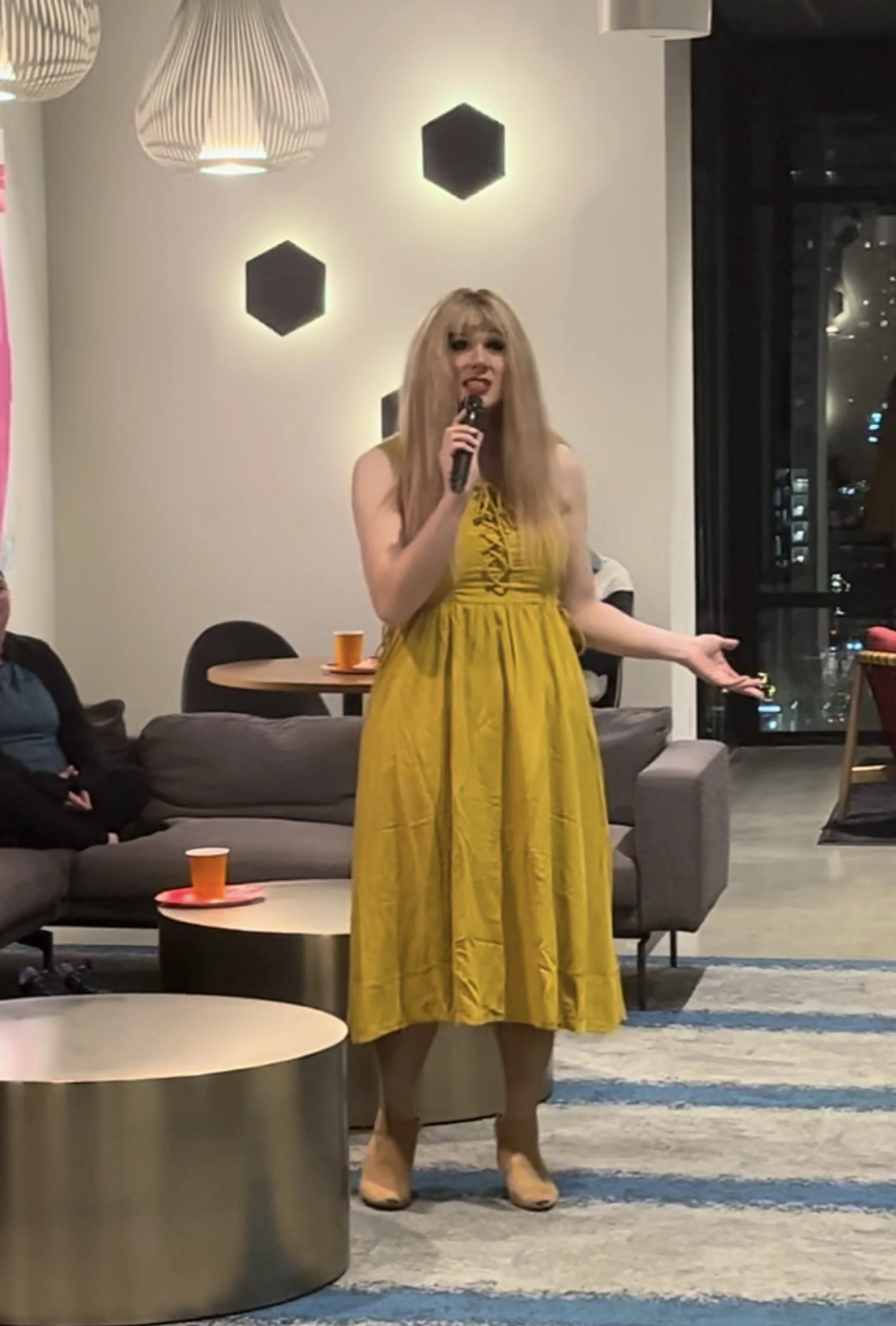 Woman in a yellow dress speaking into a microphone in a modern living room with black hexagonal wall decorations, a large window, and a gray sofa with orange cups on small tables.