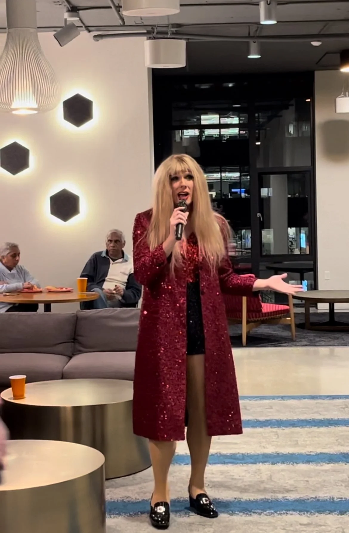 A person with long blonde hair and bangs singing into a microphone in a modern, well-lit room. They are wearing a red, sequined coat, black dress, and black shoes. In the background, two people are sitting at a table, and some wall and ceiling lights