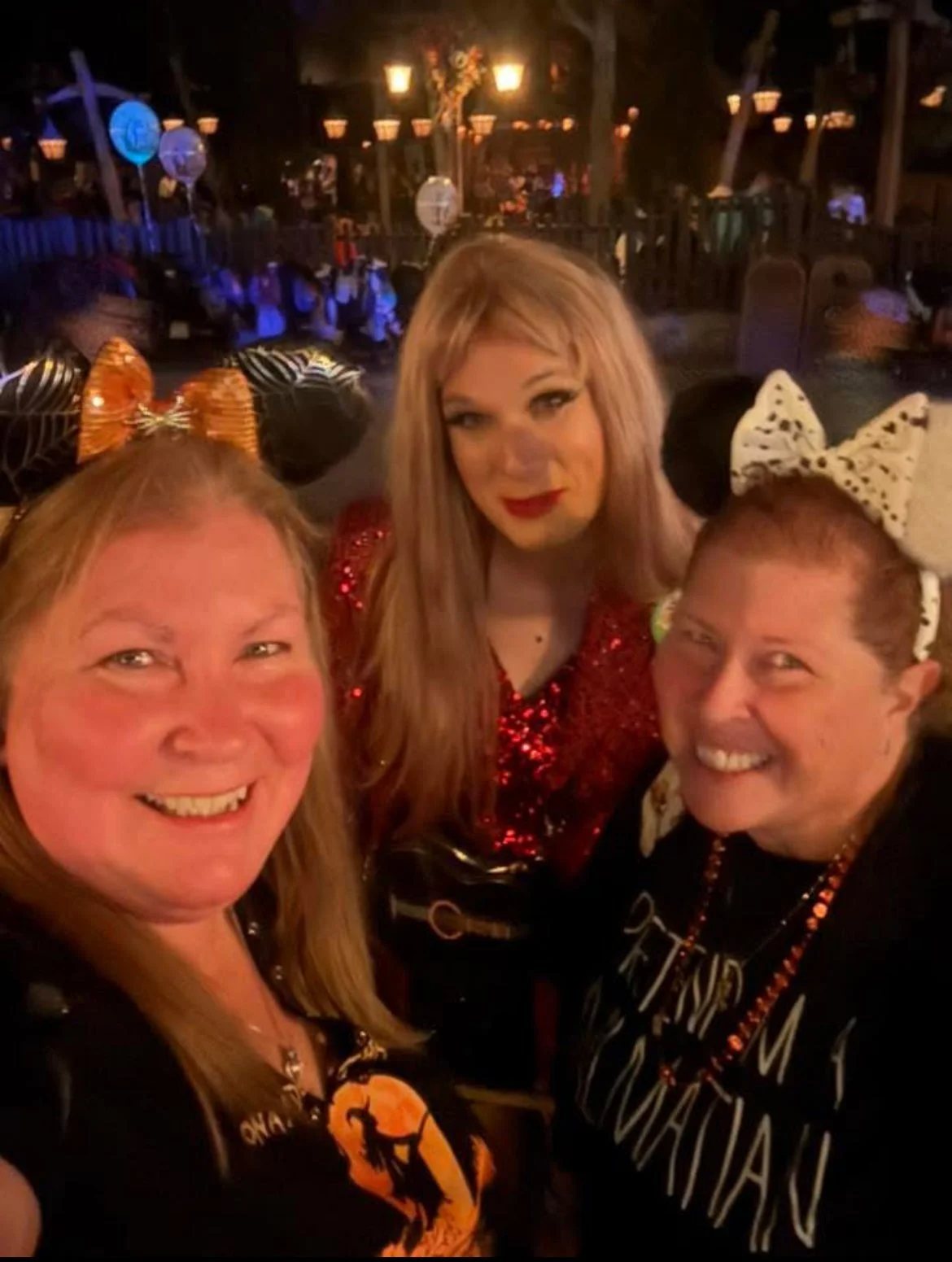 Three women smiling and taking a selfie in a festive, Halloween-themed party setting with dark lighting and decorative lanterns in the background. They are wearing Halloween costumes and accessories, including headbands with bows and ears.