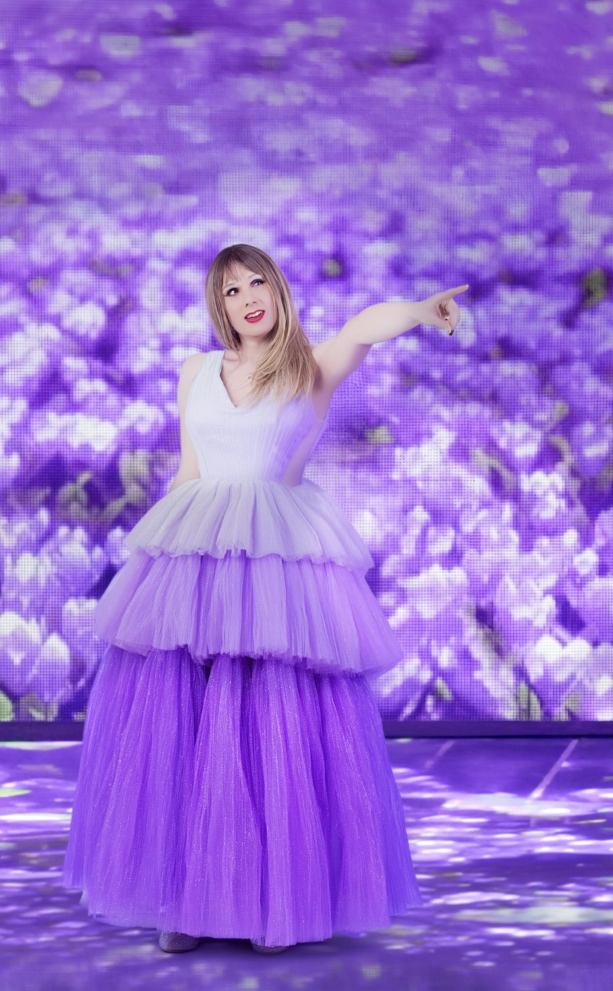 A woman wearing a lavender tutu dress is pointing to the right while standing in front of a purple floral background.