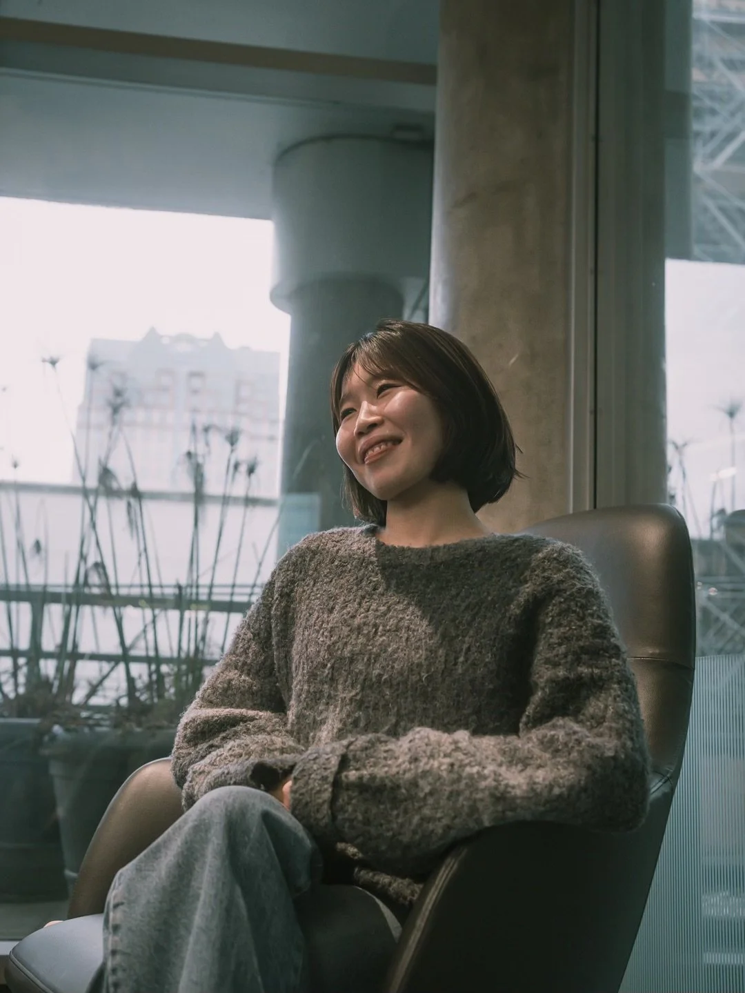 A woman with short dark hair sitting in a modern office, smiling and looking to the side, with city buildings and plants visible through large windows behind her.