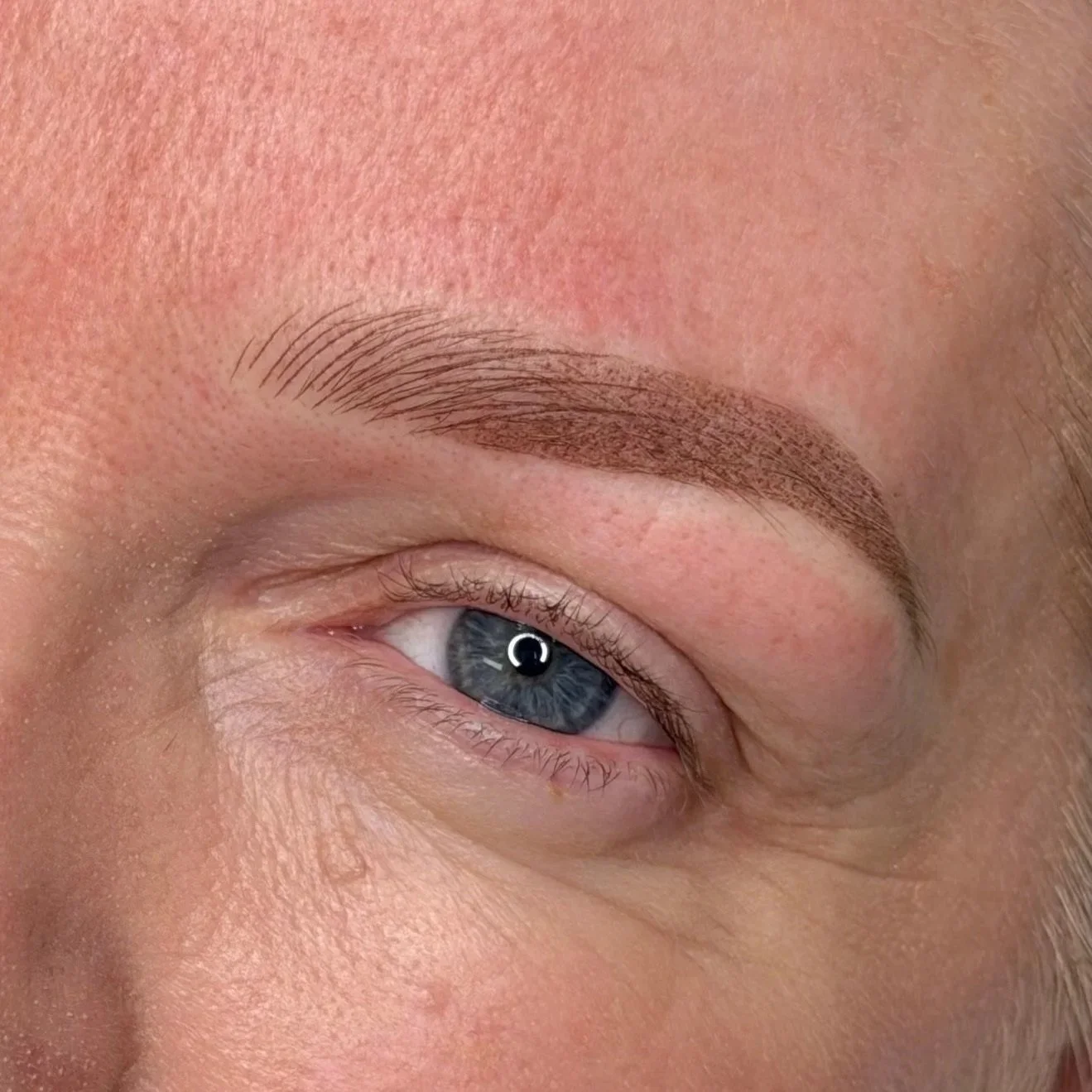 Close-up of a person's eye with blue iris, and a freshly filled thin brow tattooed with pigment.
