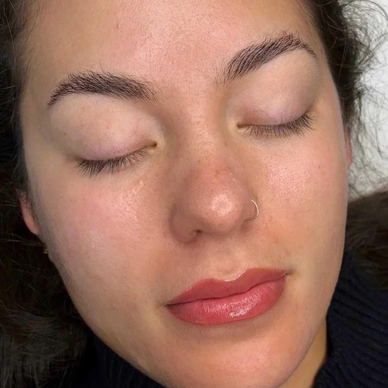 close-up of a woman's face with her eyes closed, showing her eyebrows, nose with a nose ring, and lips.