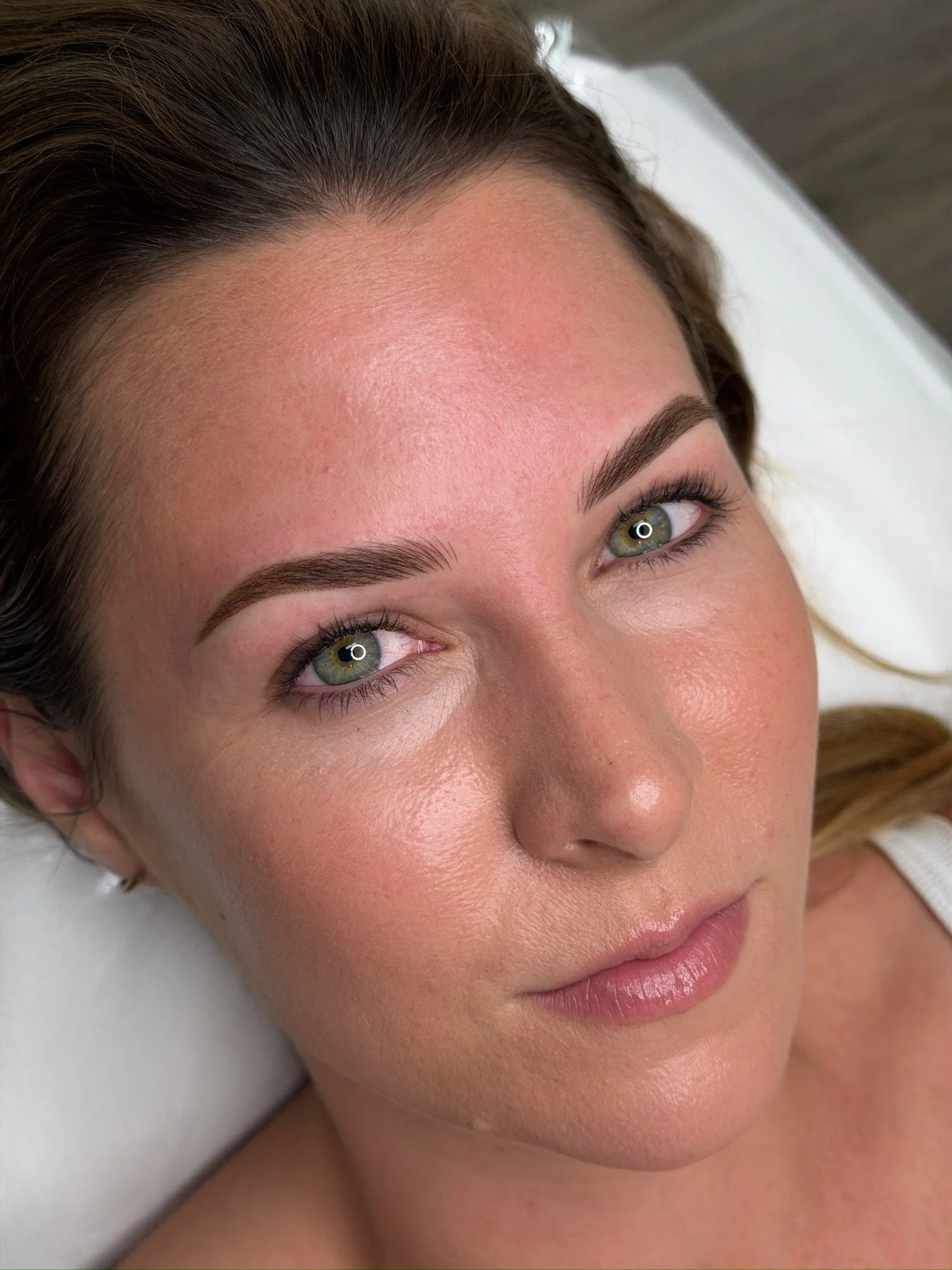 Close-up of a woman's face showing her forehead, green eyes, eyebrows, and lips, with smooth skin and natural makeup, lying on a white surface.