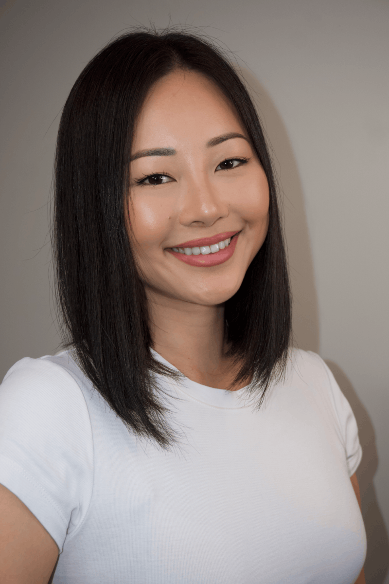 A young woman with straight, shoulder-length black hair, smiling and wearing a white t-shirt, standing against a neutral background.