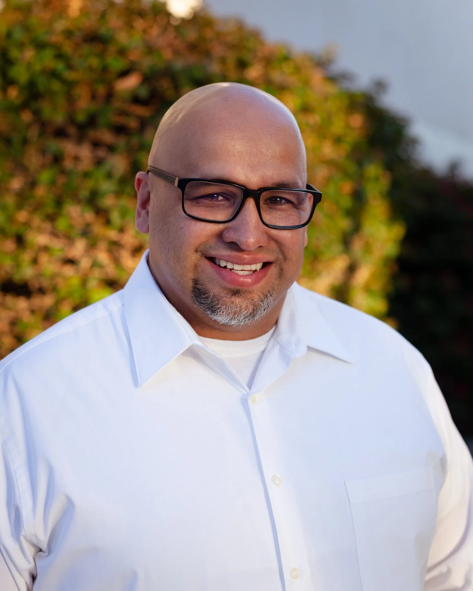 A man with a shaved head, glasses, and a goatee smiling in front of a background of trees with autumn leaves.