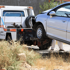 A silver sedan car is on a tow truck, being loaded for transport in an outdoor area with grass and some trees.