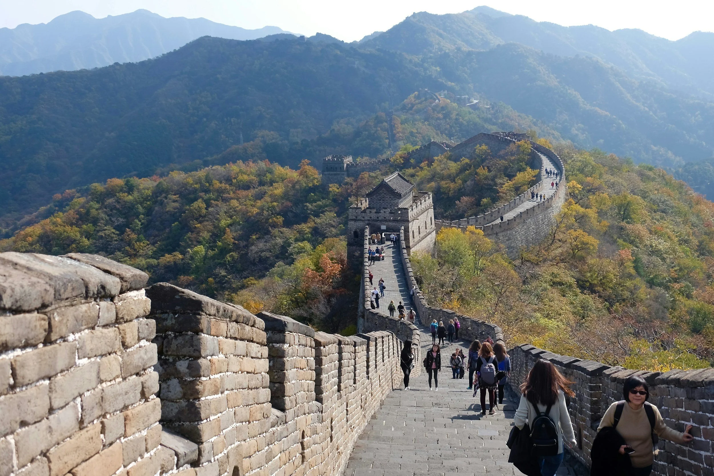 People walking on the Great Wall of China through a mountainous, wooded landscape with autumn foliage.