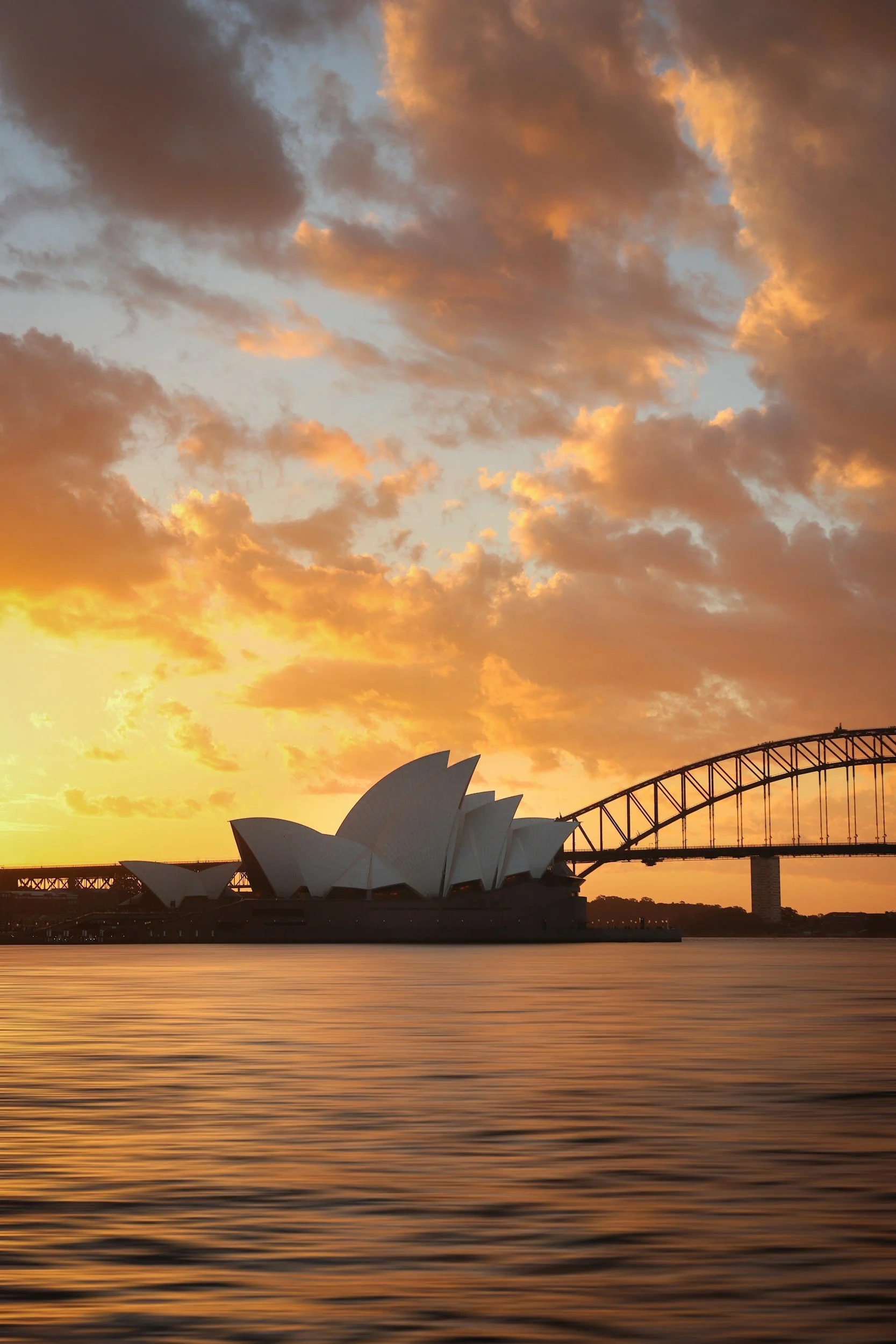 Sunset over Sydney Opera House and Harbour Bridge with colorful sky and calm water in the foreground.