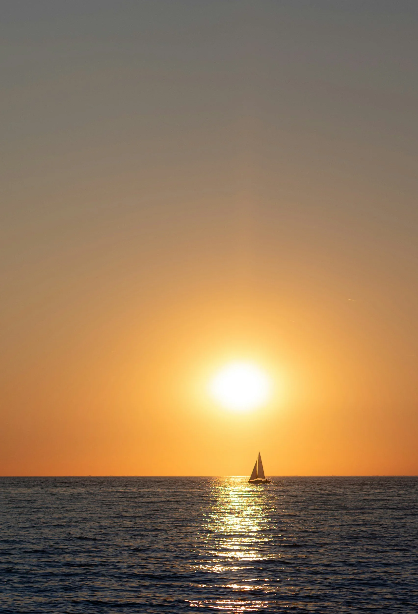 Sailboat sailing on the ocean during sunset with a bright sun and warm orange sky.