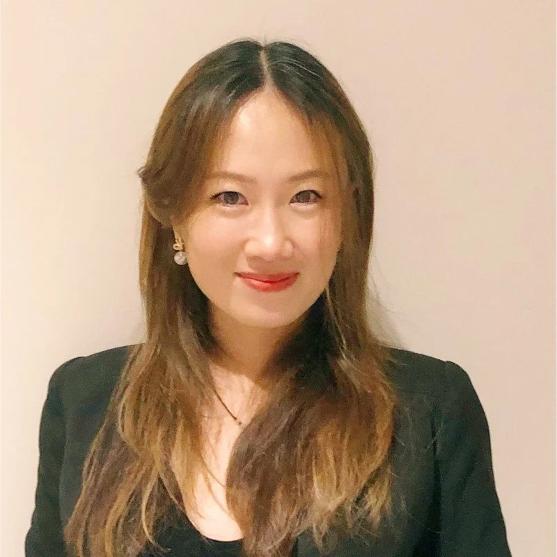 A woman with long, wavy brown hair wearing a black blazer and earrings, smiling at the camera against a plain off-white background.