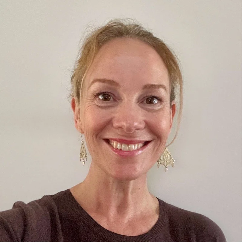 A woman with short, curly blonde hair smiling at the camera, wearing earrings and a dark brown top, standing against a plain white background.