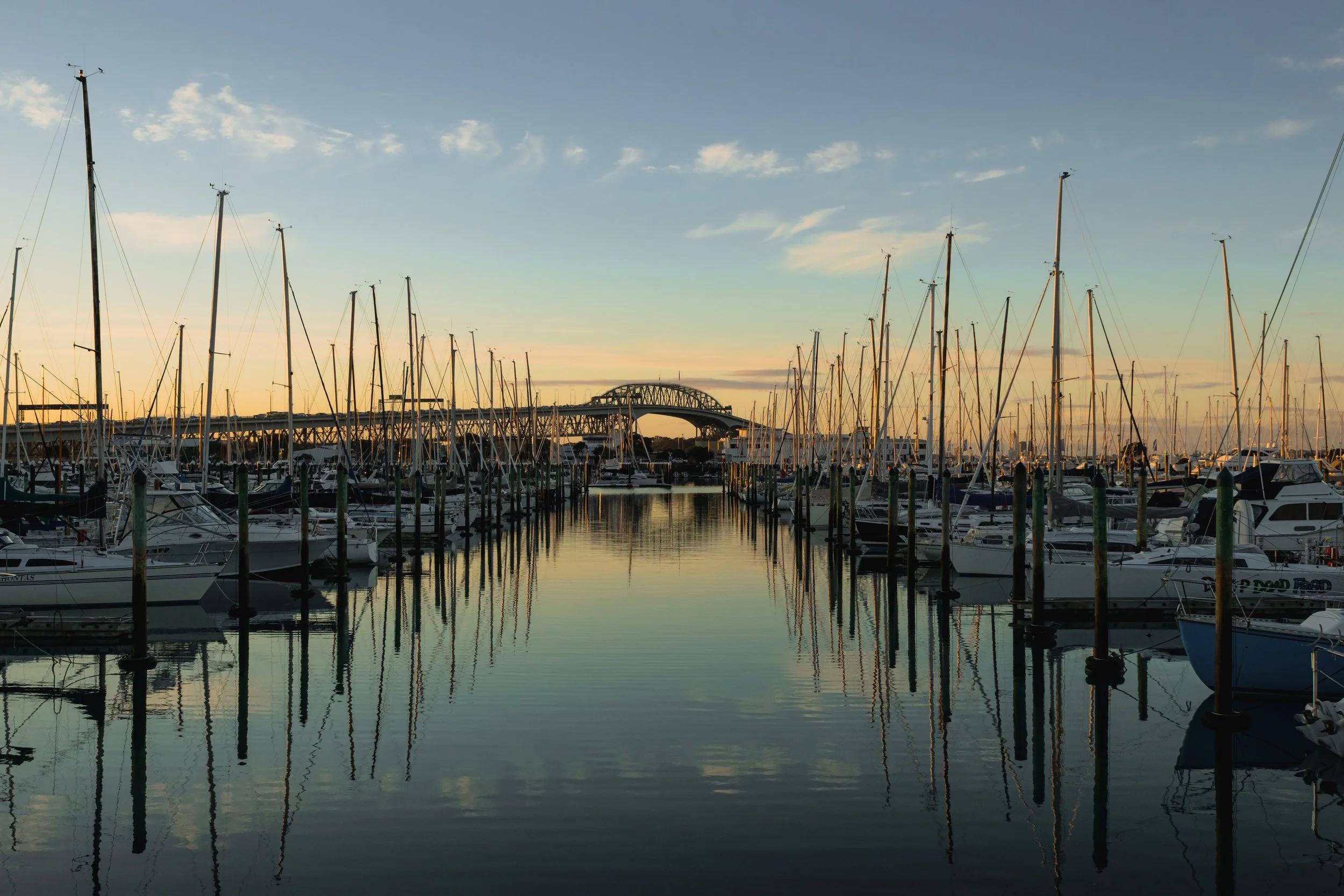 A marina at sunset with sailboats docked and a bridge in the background under a partly cloudy sky.