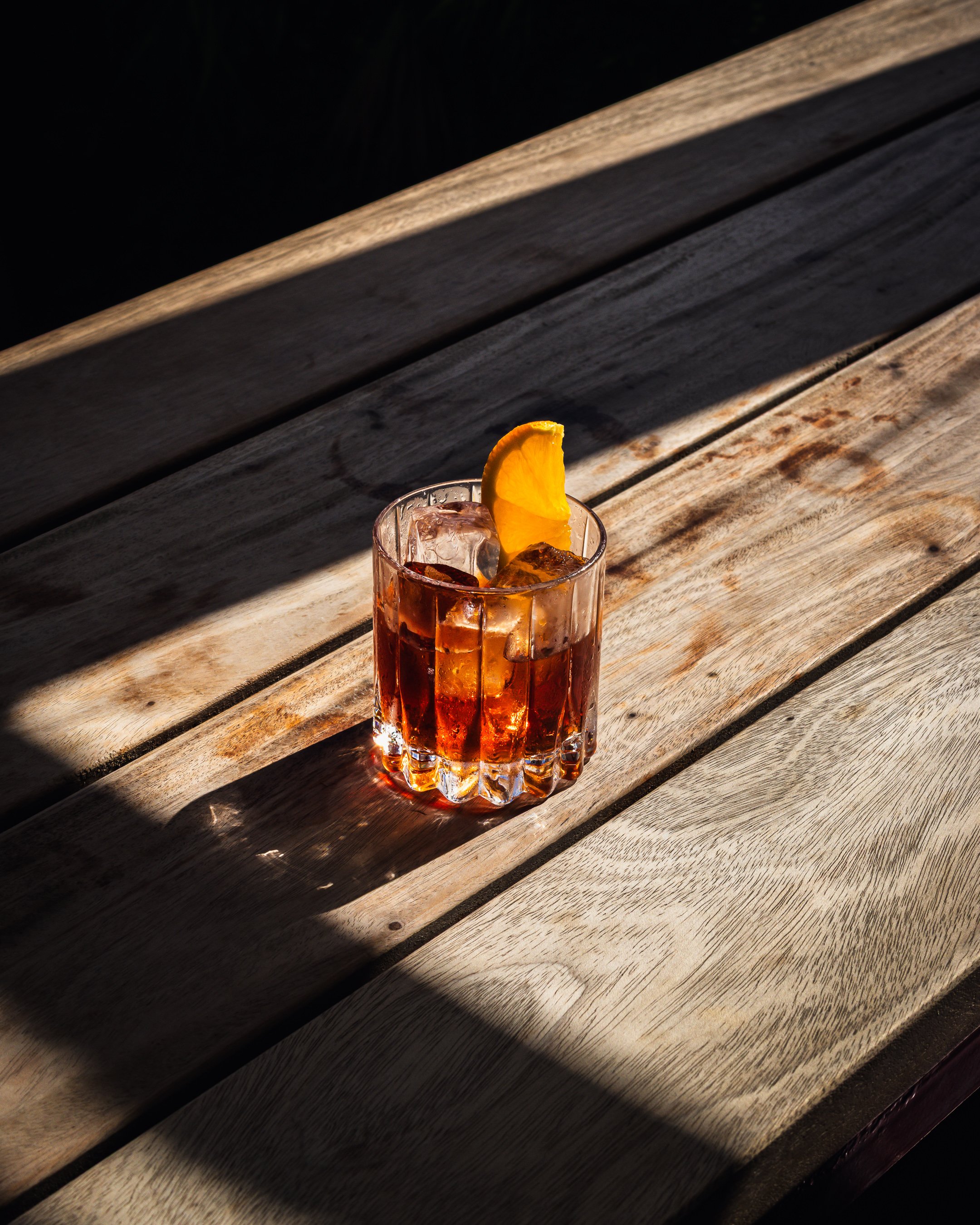 A Negroni cocktail sitting in the sunlight on a wooden table at sunset.