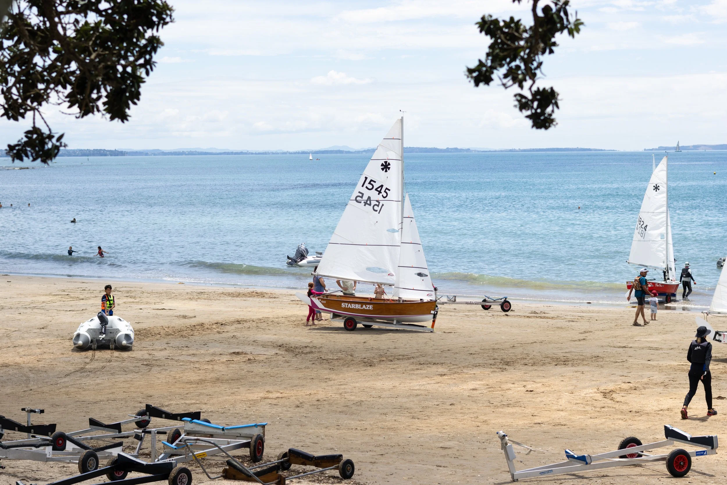A beach scene with people preparing small boats and sailboats. The ocean has sailboats in the distance, and the sky is partly cloudy. There are trees at the top edges of the image and some children and adults near the water.