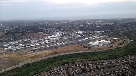 Aerial view of a large industrial area, a nearby residential neighborhood and open land with roads on the horizon under a cloudy sky.