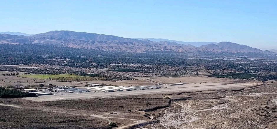 Aerial view of an arid landscape with a small industrial or commercial area, surrounded by mountains and distant city, under clear blue sky.