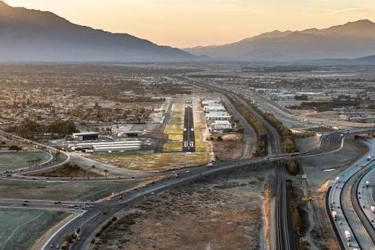 An aerial view of a desert landscape with roads, highways, and an airport runway, with mountains in the background at sunset.