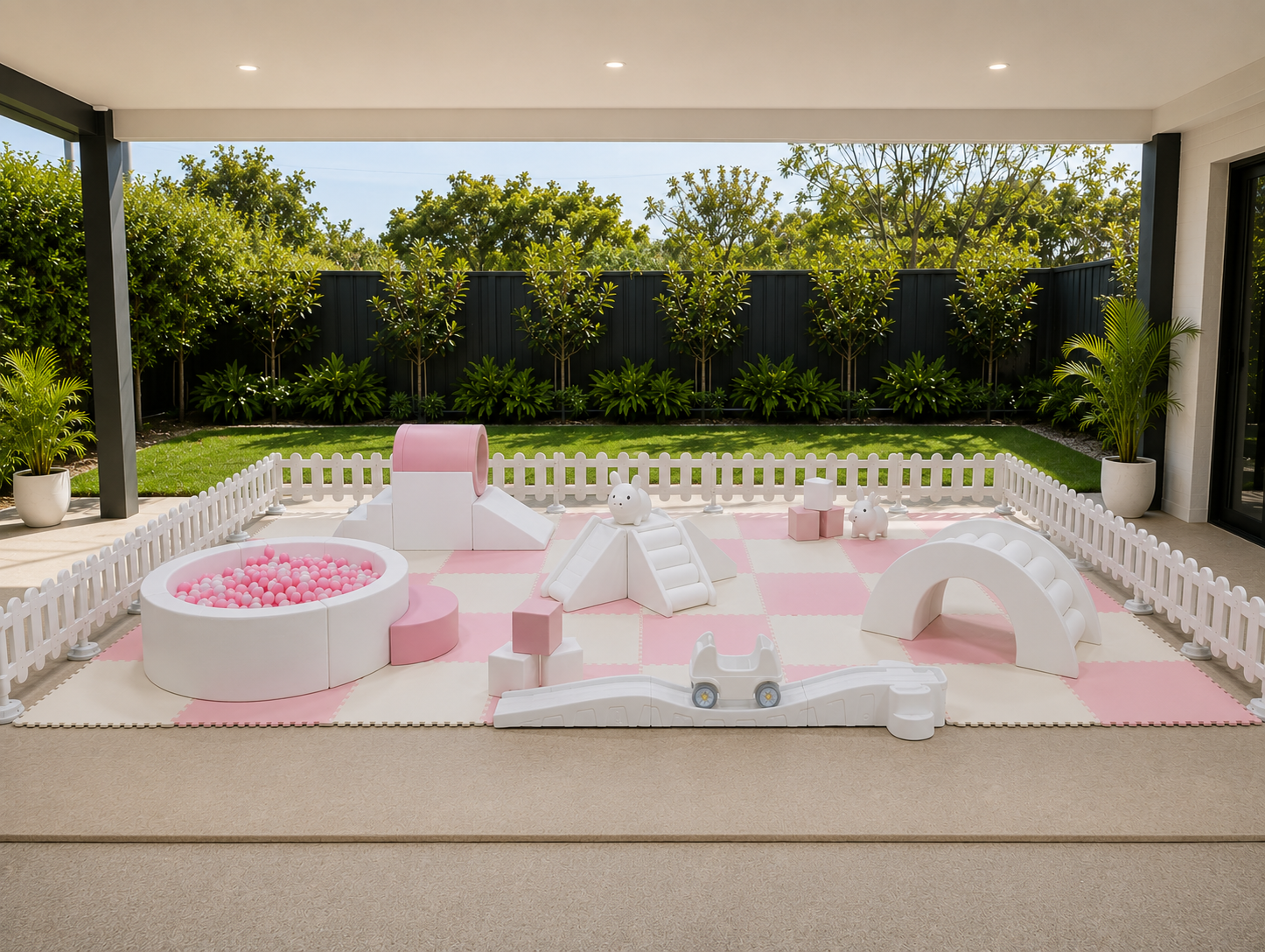 Indoor play area with pink and white toys, ball pit, climbing structures, and a small train on a foam tile floor, enclosed with a white picket fence, overlooking a green backyard with trees.