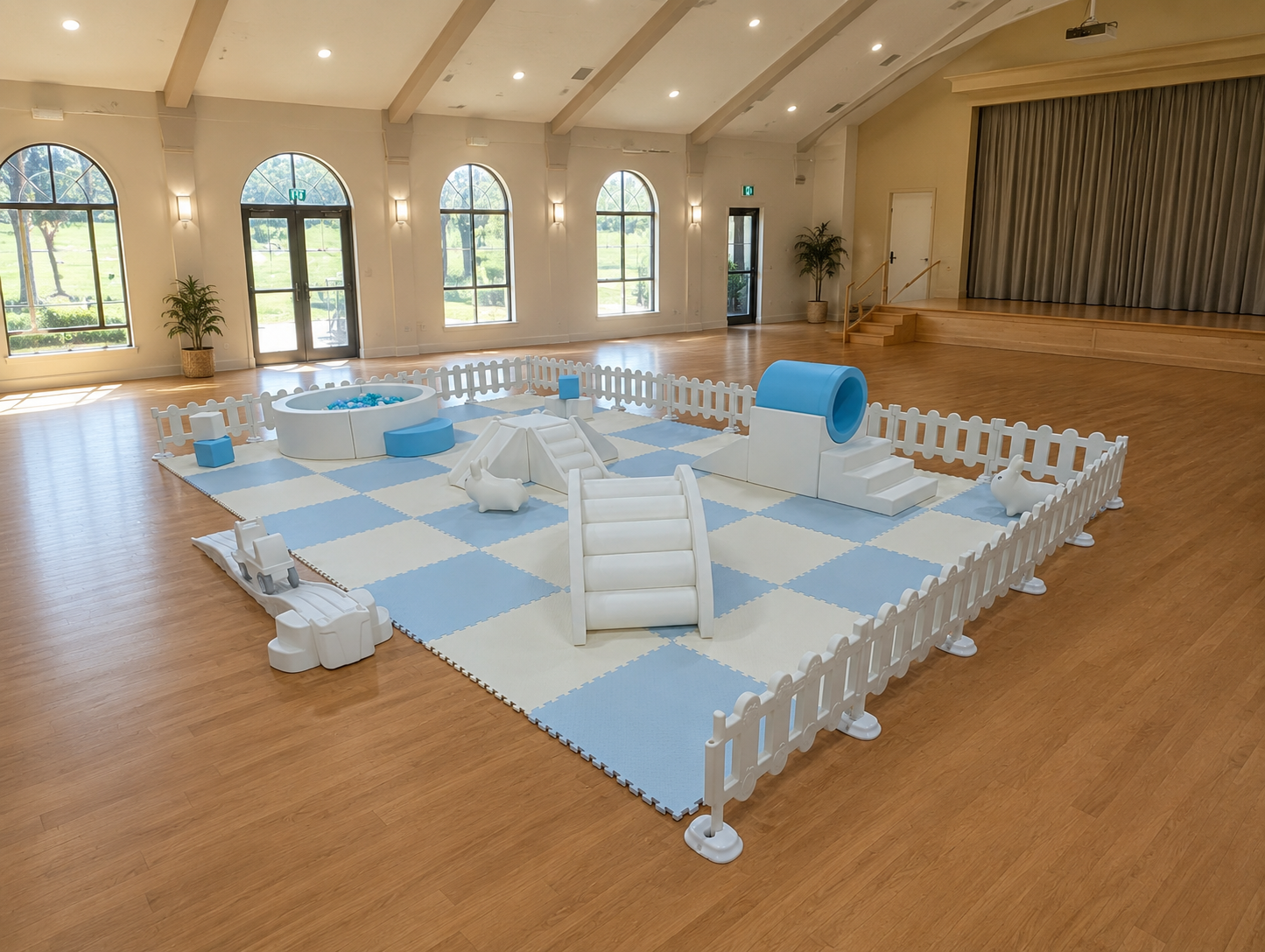 Empty indoor play area with soft blue and white checkered padded flooring, small plastic play structures including a slide, tunnel, and animal figures, surrounded by a white picket fence, in a bright room with large arched windows and wooden floors.
