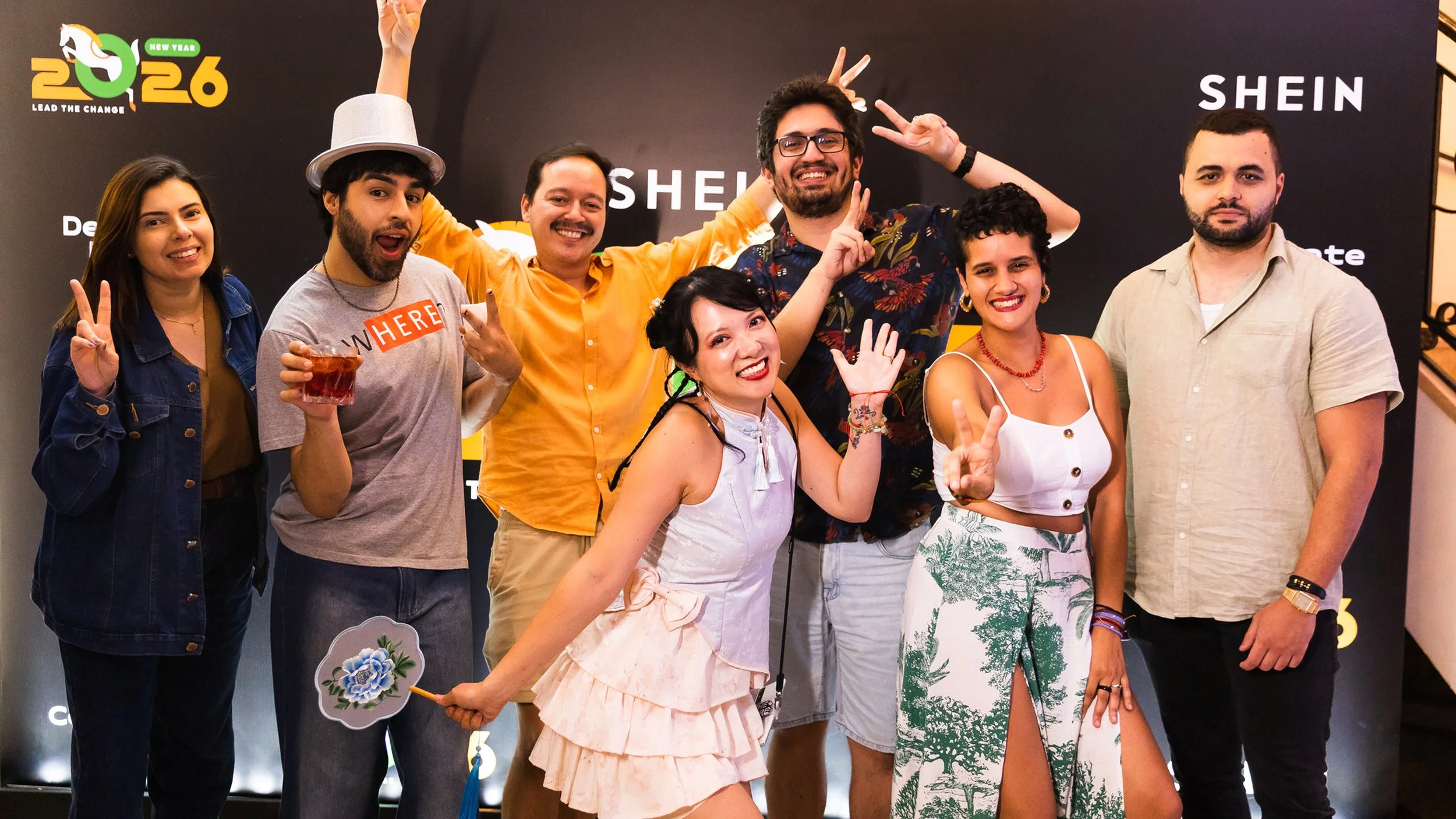 Group of eight friends smiling and posing together at an event, some making peace signs, with a black backdrop featuring the logos 'SHEIN' and 'Lead the Change 2026' in the background.