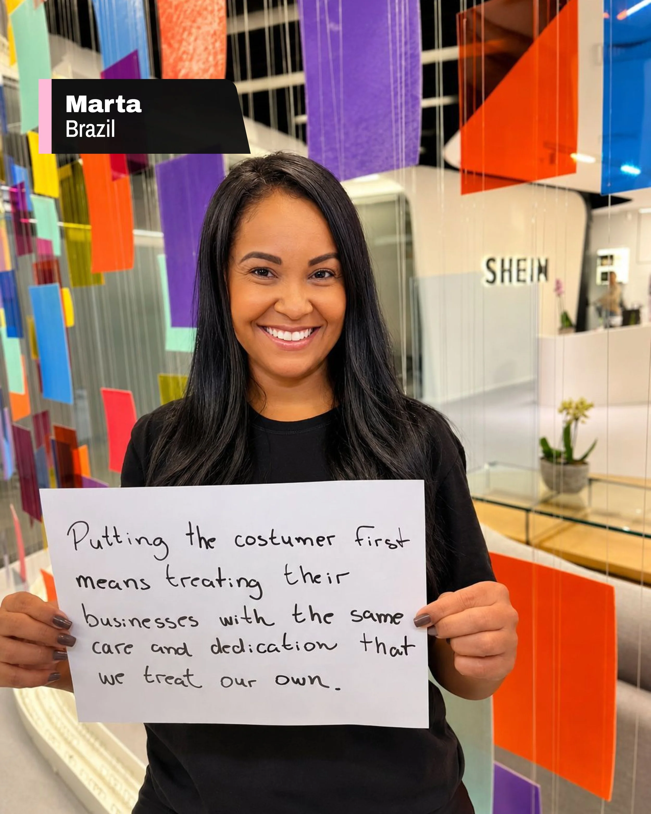 A woman with long dark hair smiling, holding a handwritten sign, standing in front of colorful hanging fabric or paper decorations in a store. The sign discusses treating animals with care and dedication.