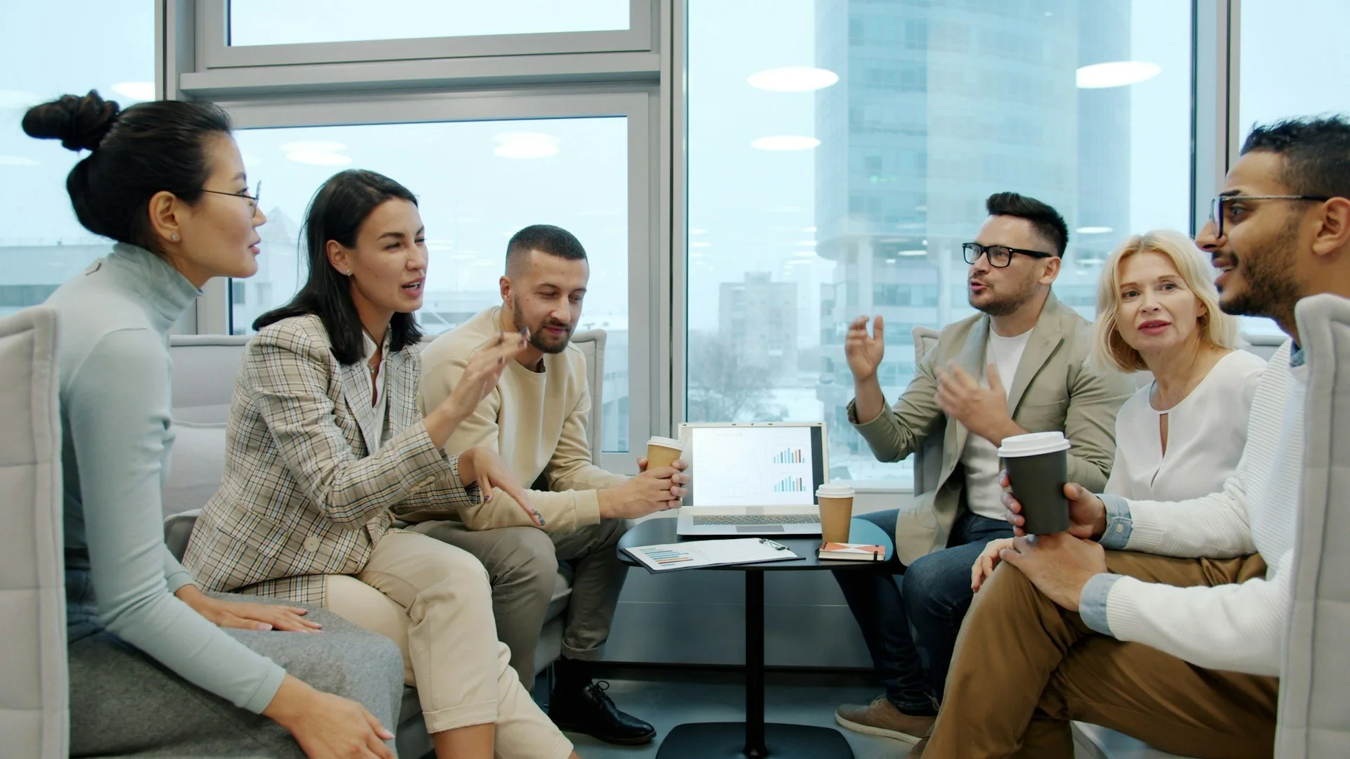 Group of six diverse people having a discussion in a modern office with large windows and city view, using laptops and coffee cups.