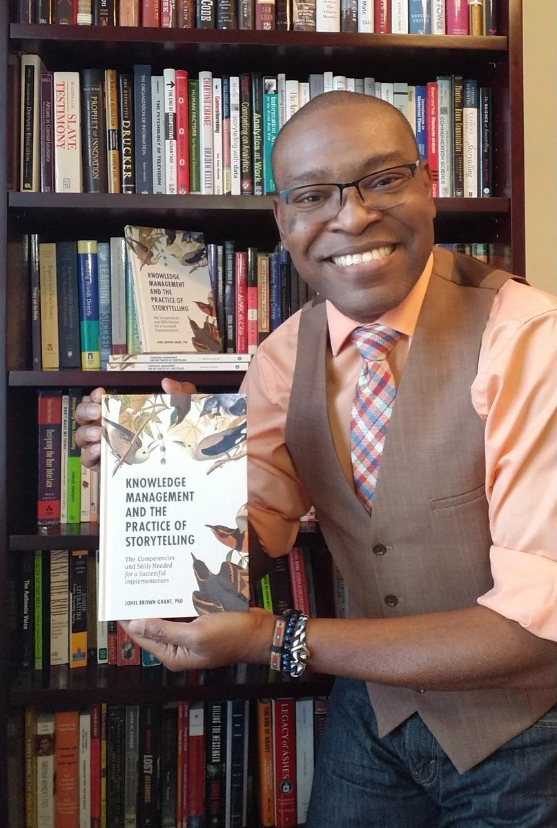 A smiling man with glasses, dressed in a peach shirt, brown vest, and colorful tie, holding a book titled "Knowledge Management and the Practice of Storytelling" in front of a bookshelf filled with various books.