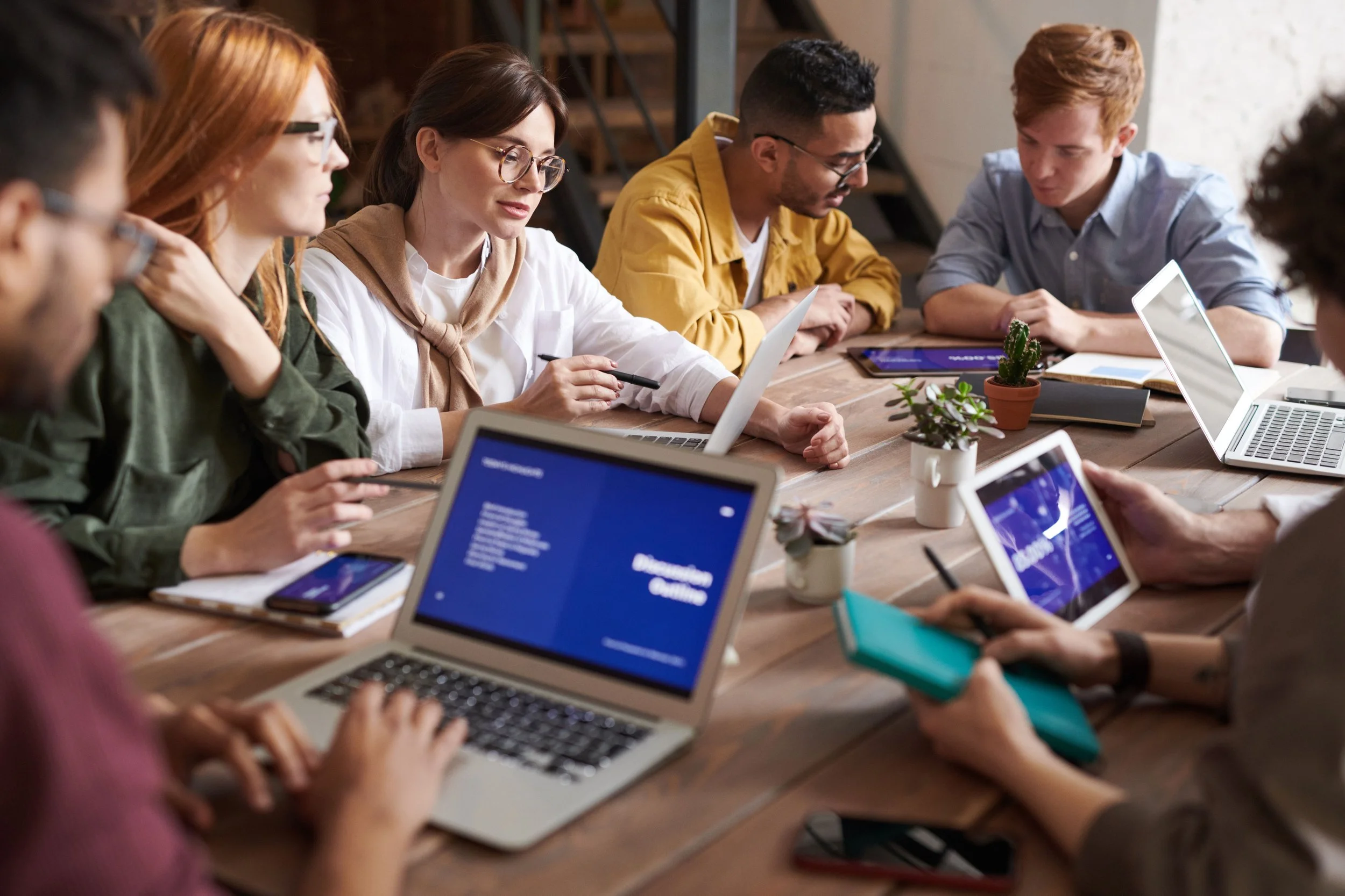 A group of diverse young adults sitting around a wooden table in a meeting room, engaged with laptops, tablets, and notebooks, discussing and collaborating.
