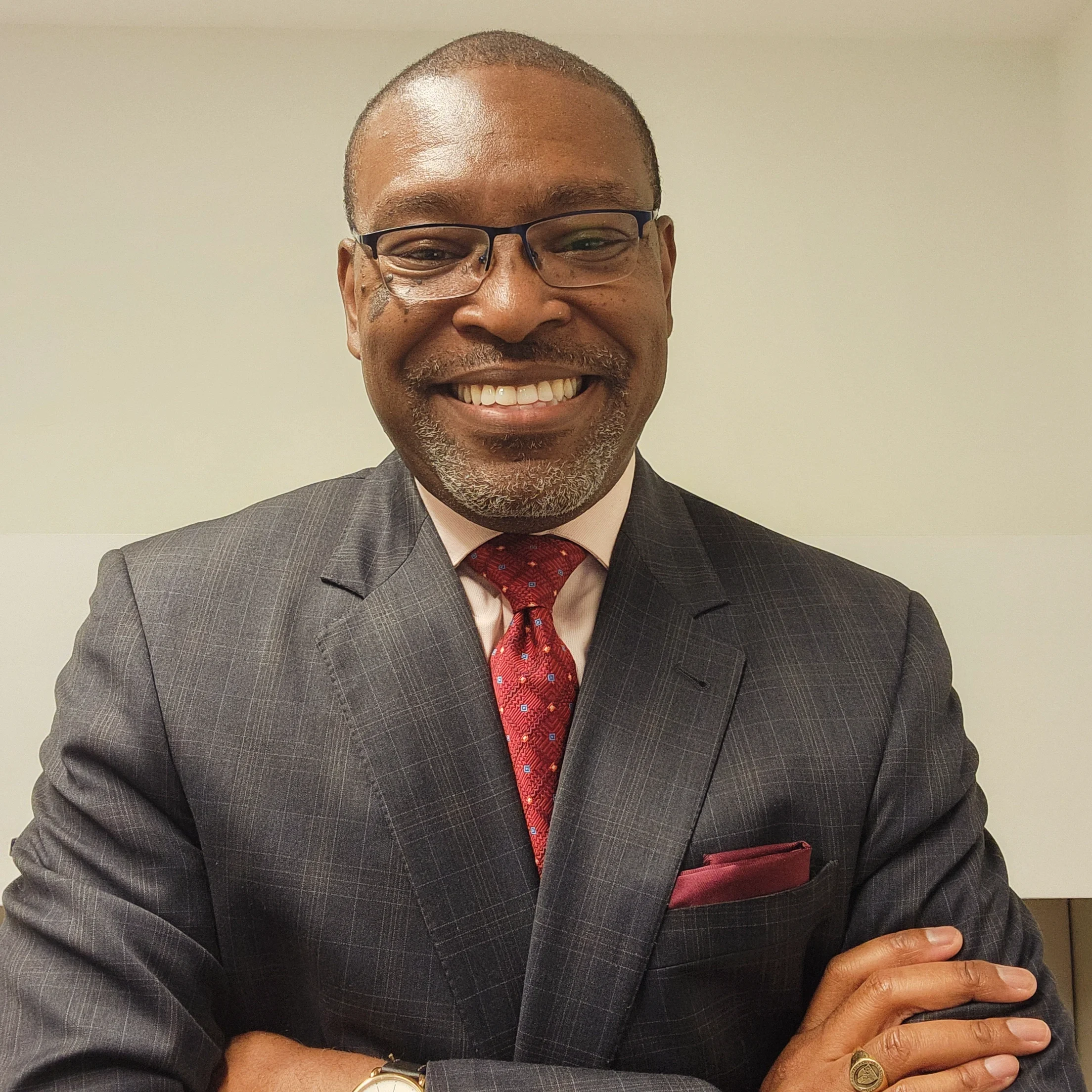 Close-up of a smiling middle-aged man with dark skin, wearing glasses, a business suit with a checkered pattern, a pink shirt, a red patterned tie, and a matching pocket square. He is also wearing a gold watch and a ring on his finger, with a neutral light-colored background.