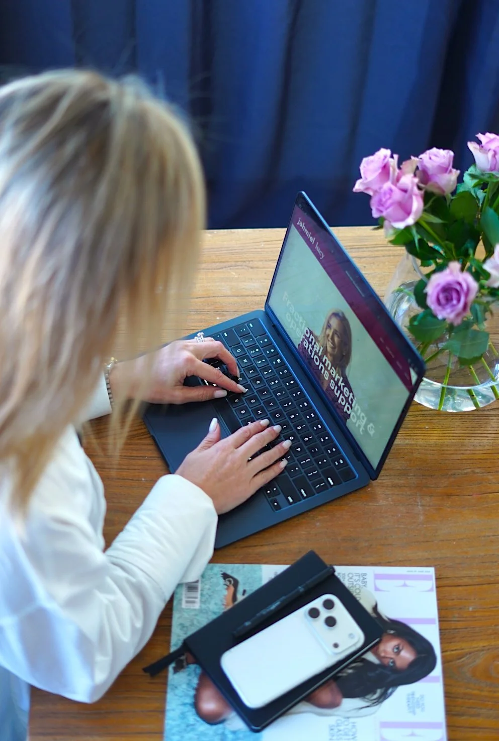 Marketing Strategy & Marketing Operations Services - Jahmiel Lucy. A woman with blonde hair using a laptop on a desk with a smartphone, magazine, and a vase of pink roses nearby.