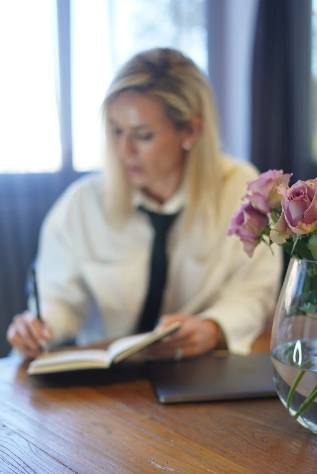 Marketing Strategy & Marketing Operations Services - Jahmiel Lucy. A woman with blonde hair, wearing a white shirt and black tie, sitting at a desk taking notes. A vase with pink roses is in the foreground.