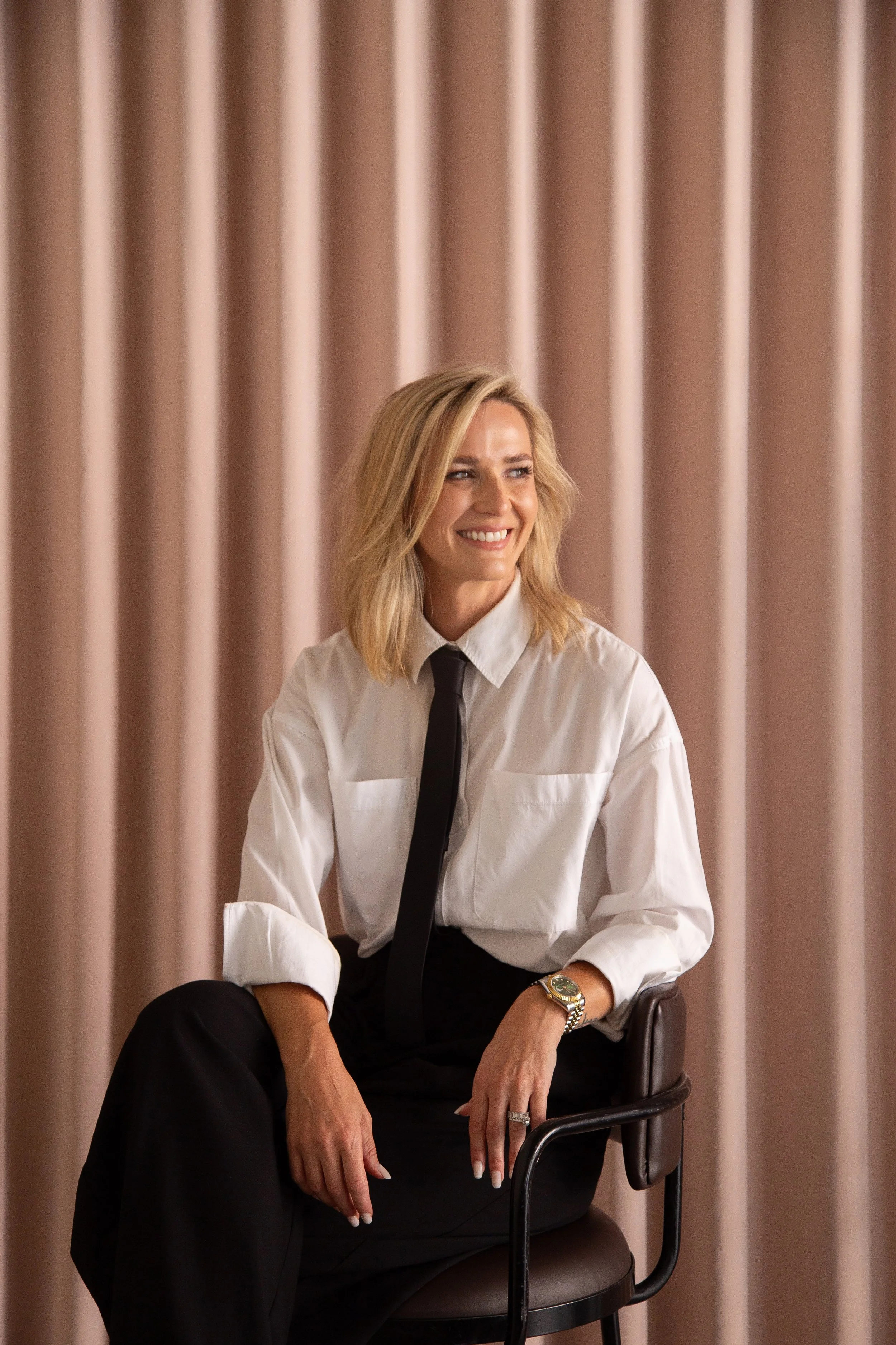 A woman with blonde hair in a white shirt, black tie, and black pants sitting on a black chair with a pink curtain in the background. Jahmiel Lucy, fractional marketing and operations consultant, Brisbane and South East Queensland