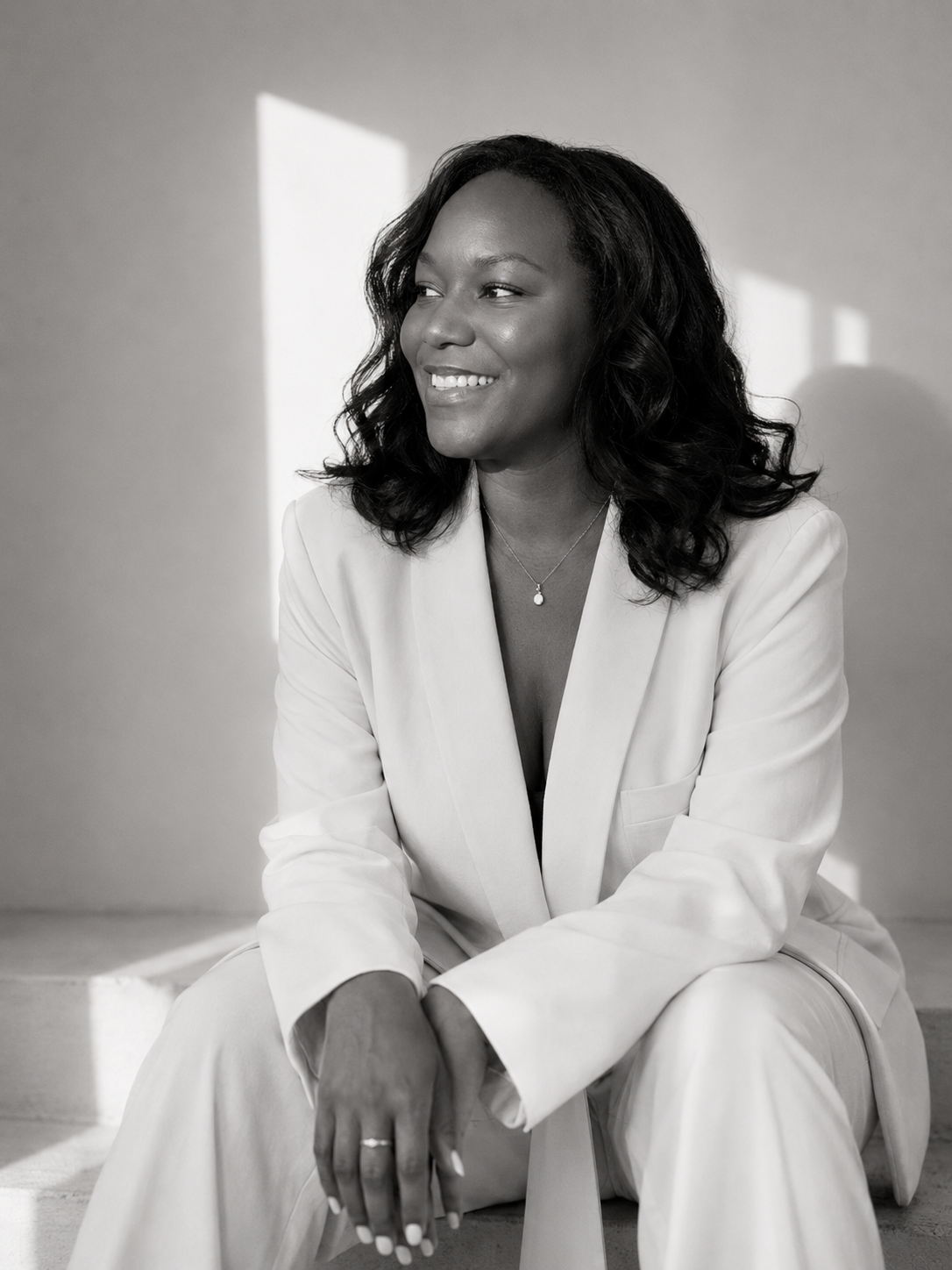 Black and white portrait of a woman in a light-colored suit sitting on the floor, smiling and looking to her left. She has wavy hair and wears a necklace and rings.