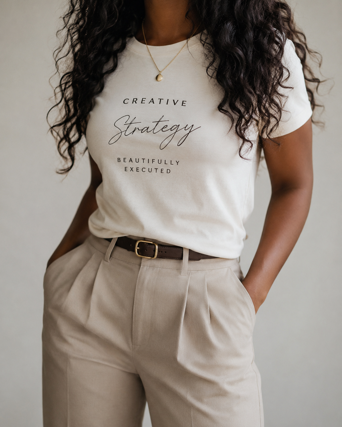 A woman wearing a white T-shirt with the words 'Creative Strategy Beautifully Executed' printed on it, tucked into beige high-waisted pants, with her hands in her pockets and wearing a gold necklace.