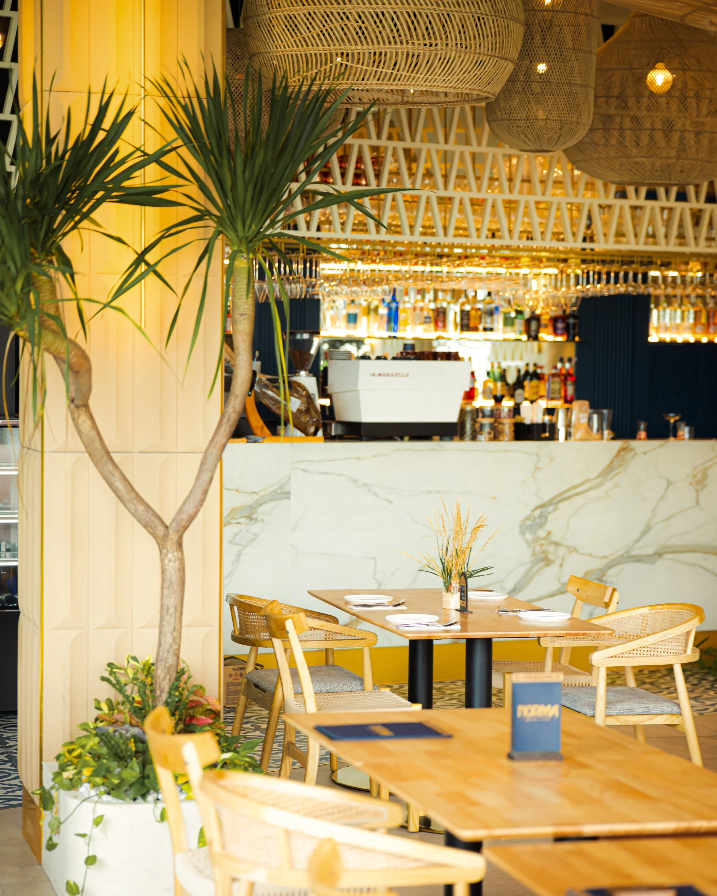 Interior of a restaurant with wooden tables and chairs, a potted plant in the foreground, and bar shelves with bottles in the background. Overhead wicker light fixtures and a marble counter are also visible.