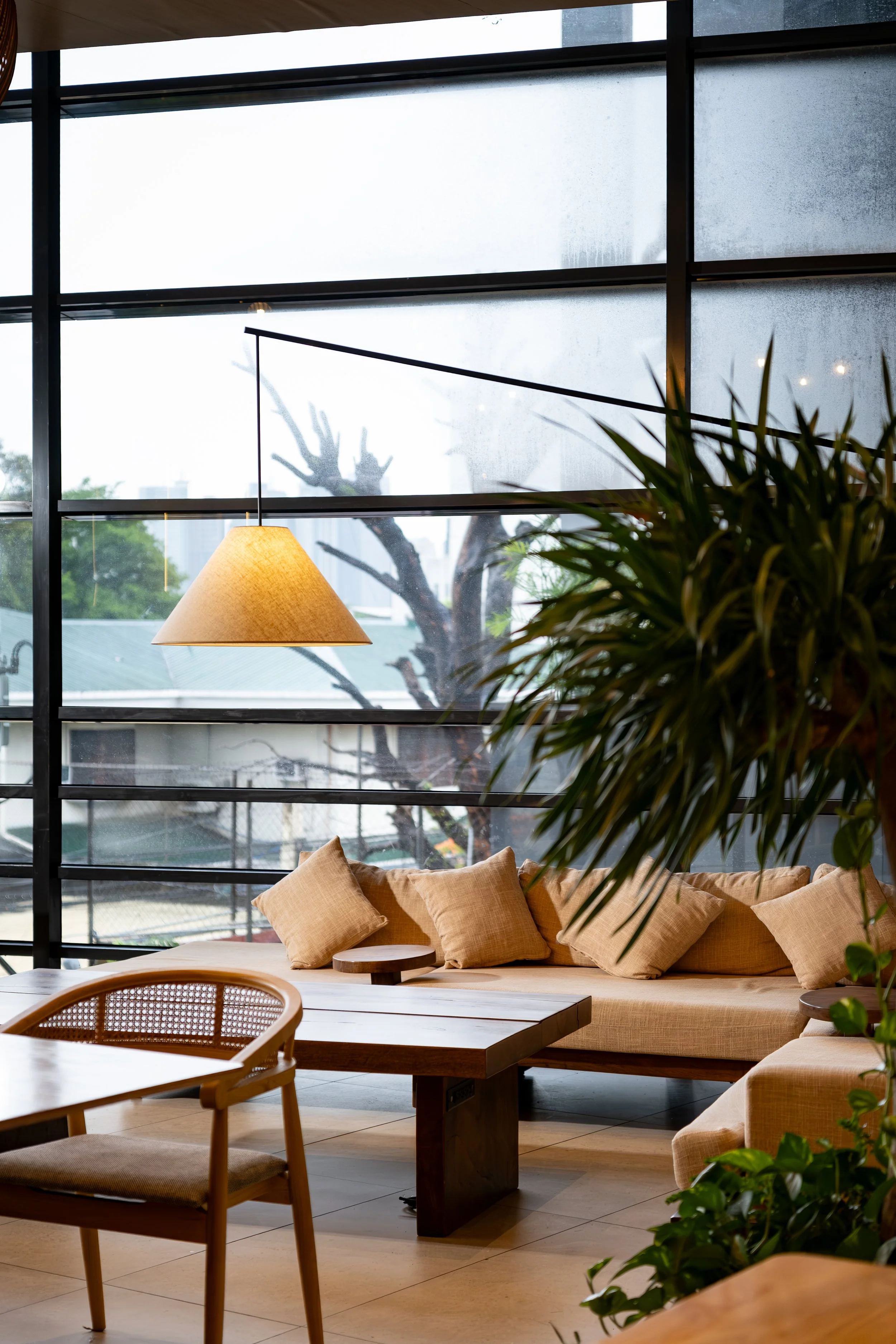 Interior of a modern lounge area with large windows, beige cushions on a built-in sofa, a hanging lamp, a wooden dining table with chairs, and lush green plants. Outside, trees and buildings are visible through the frosted and clear glass panes.