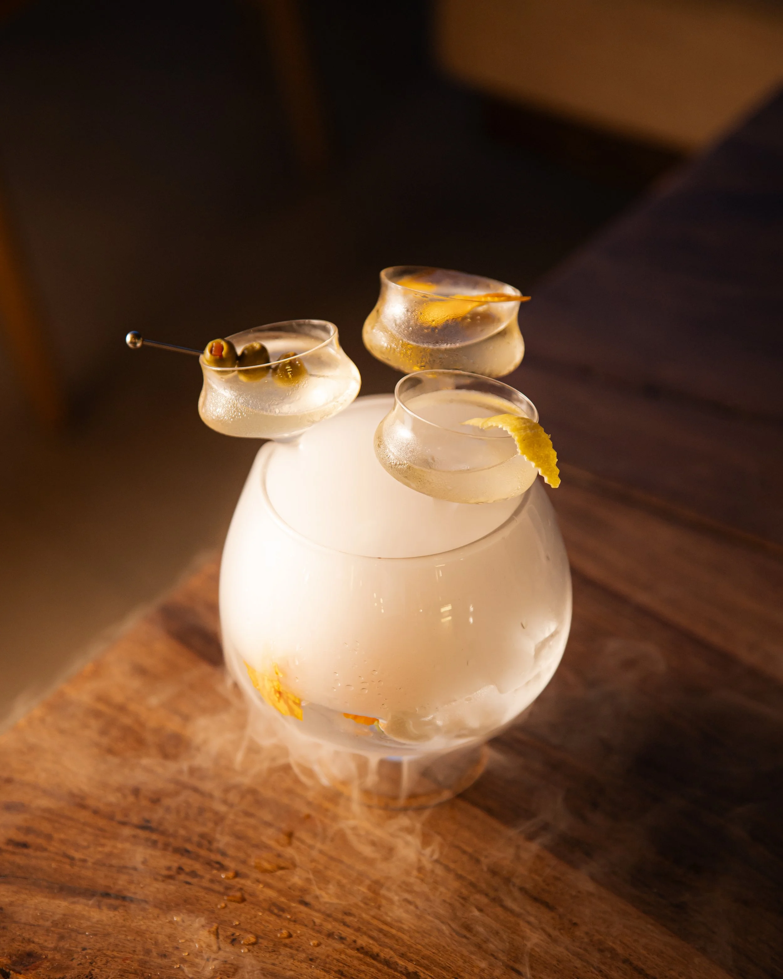 Three martini glasses on a white ceramic stand, garnished with olives, lemon twist, and citrus peel, on a wooden surface.