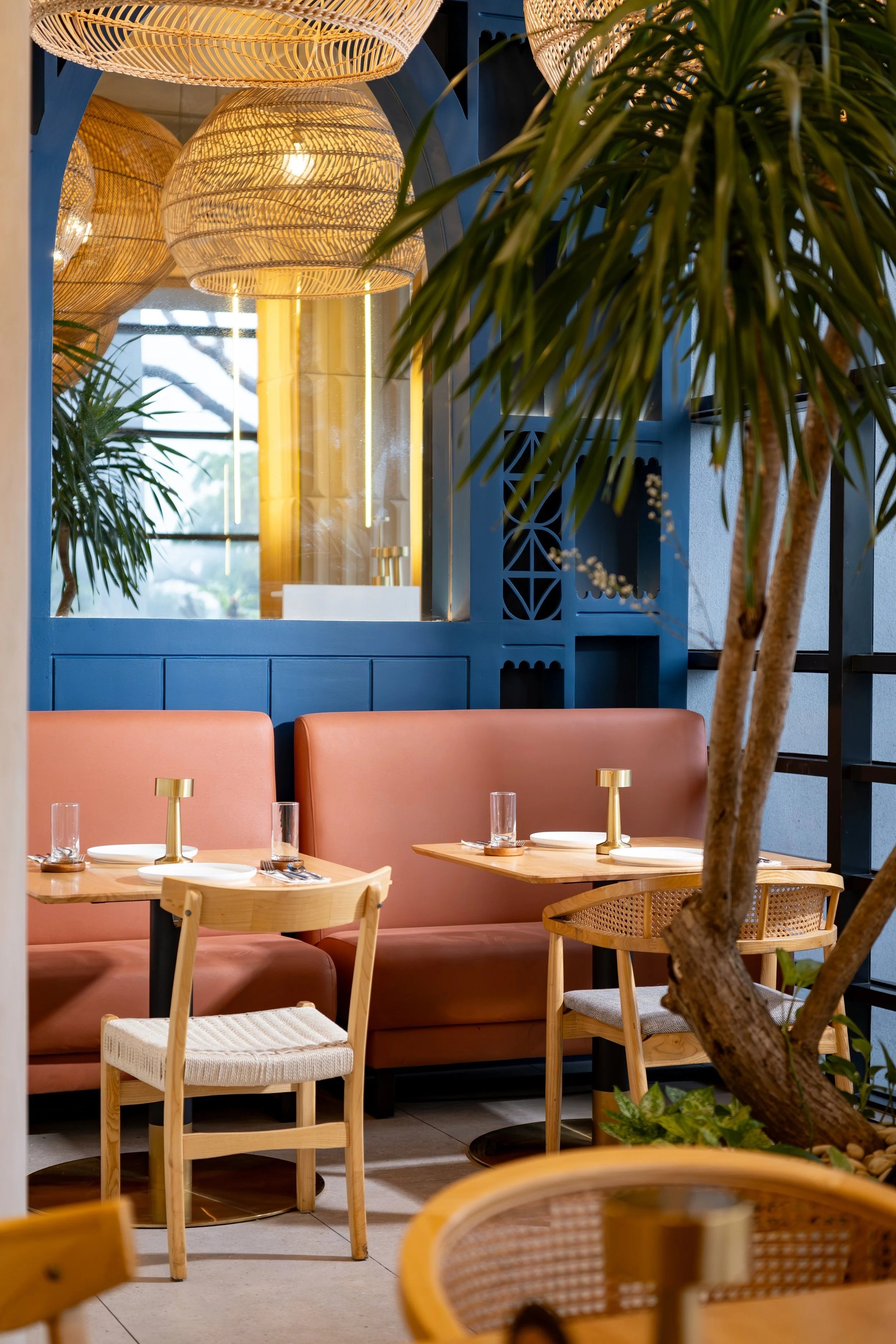 A cozy restaurant interior featuring pink padded benches, wooden chairs, small round tables set with glasses, plates, and gold candle holders. There are large woven pendant lights hanging from the ceiling, a large mirror on a blue wall, and various green indoor plants.