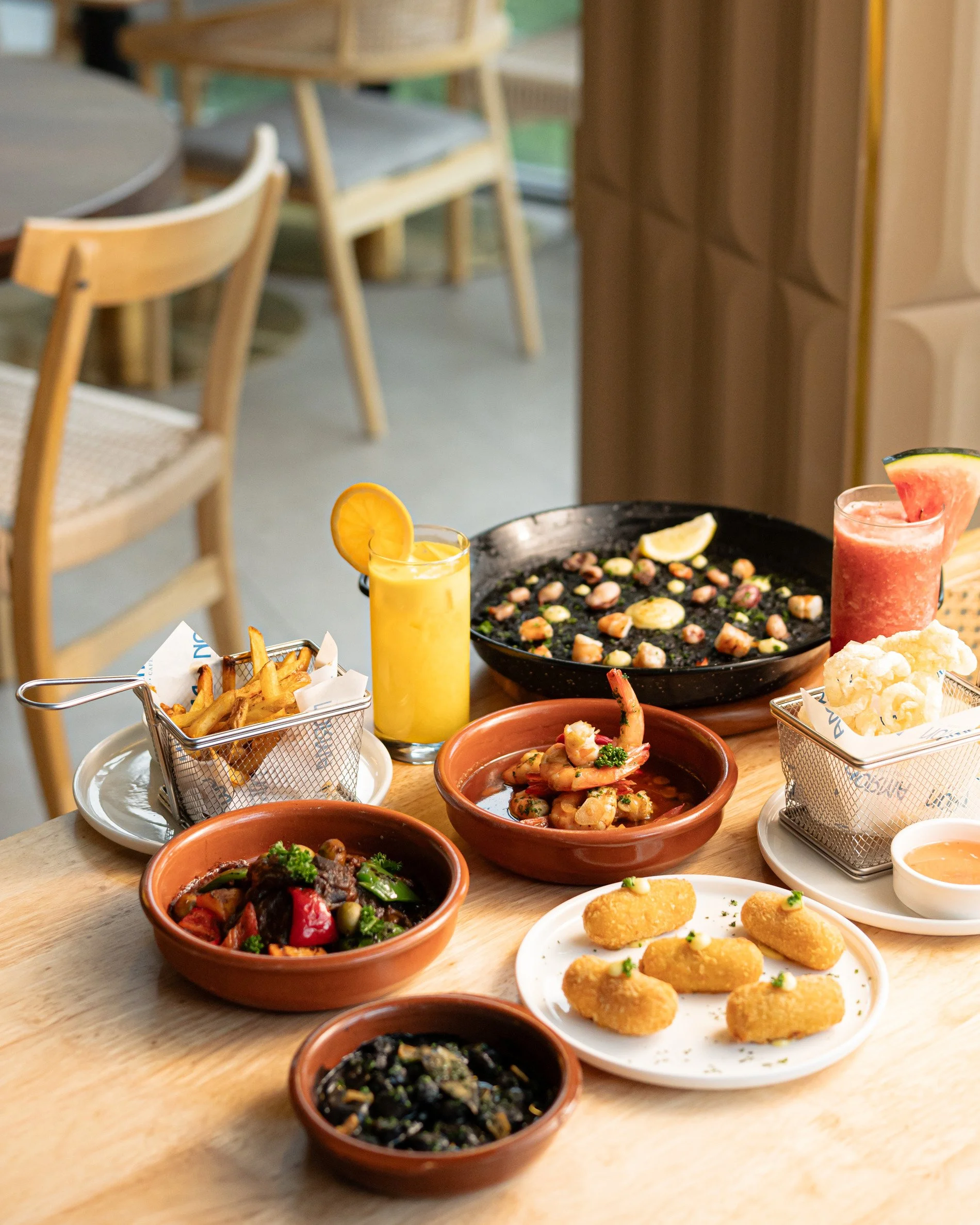 A variety of dishes on a wooden table, including bowls of salads, fried appetizers, and drinks with lemon and watermelon garnishes. The background shows chairs and a window in a cozy restaurant setting.