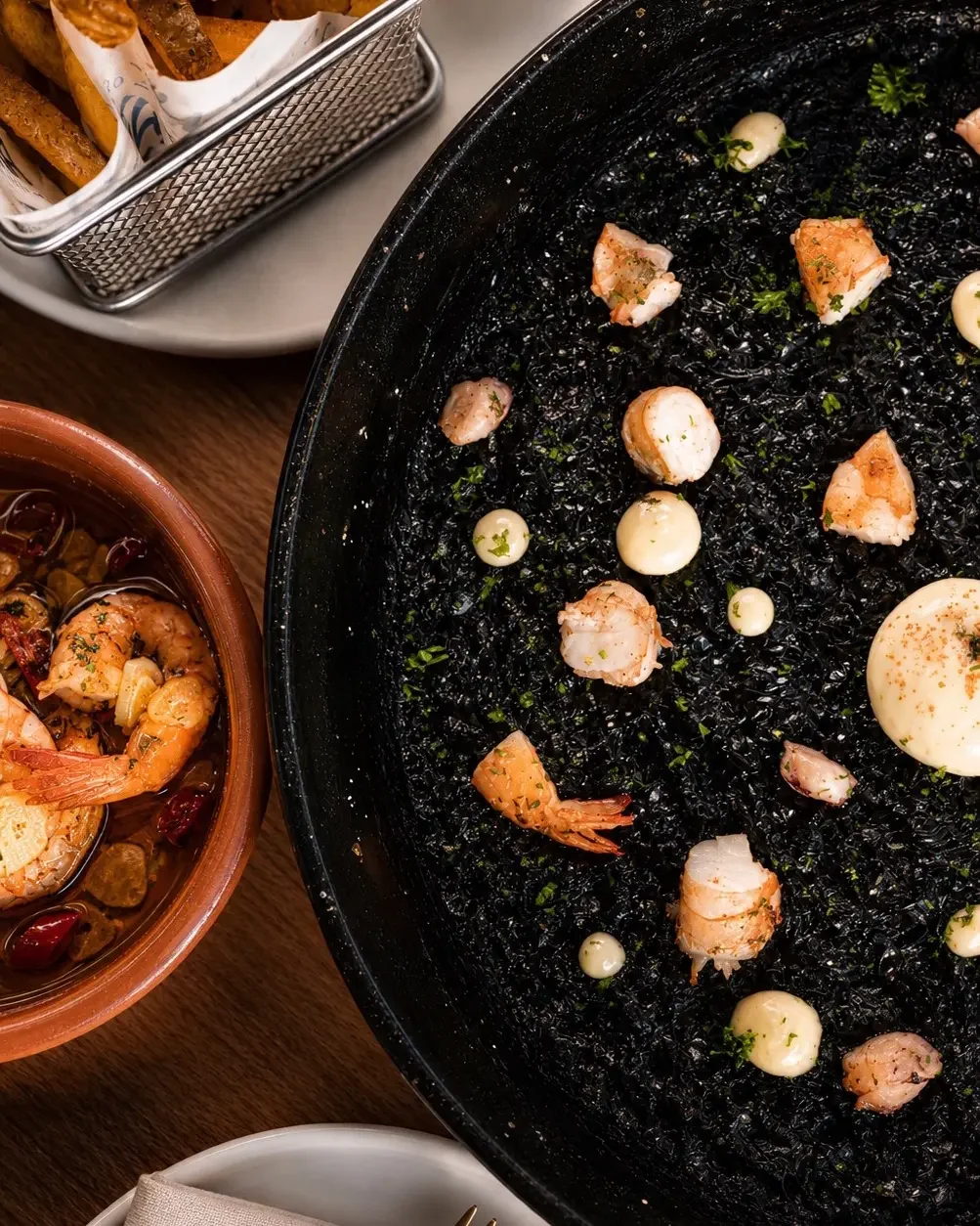 Seafood dish with shrimp and scallops cooking in a black skillet, with a side of breaded shrimp and chips in basket, and a bowl of crustacean salad with shrimp and herbs.
