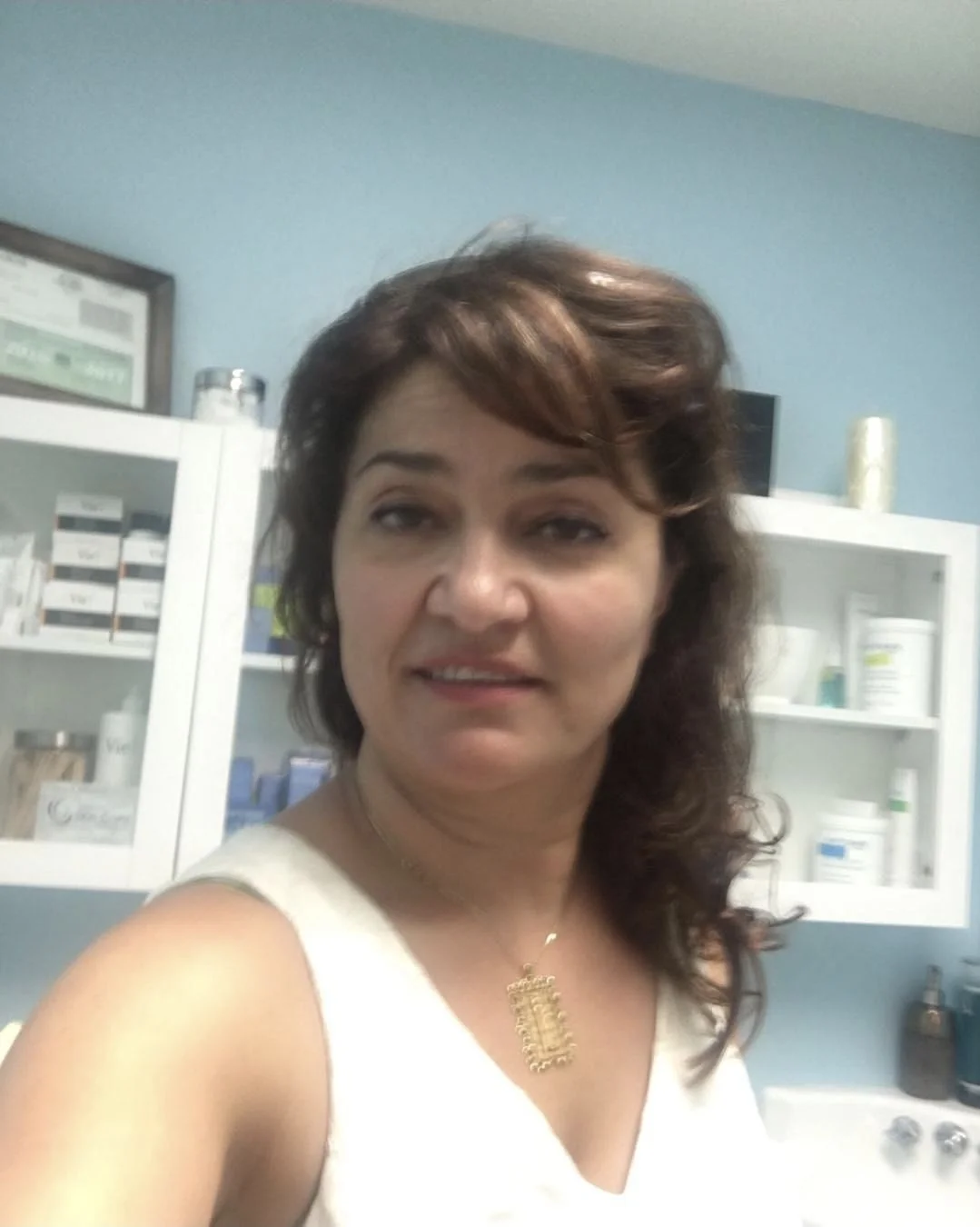 A woman with wavy brown hair taking a selfie in a room with blue walls and white cabinets containing medical supplies.