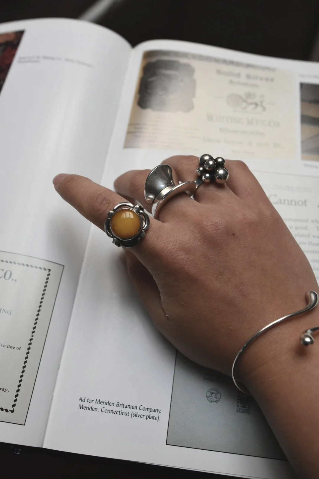 A hand with three silver rings, each with different designs and a yellow gemstone, resting on an open magazine or catalog. The hand also wears a silver bracelet with a round bead detail. The magazine contains vintage advertisements and photographs.