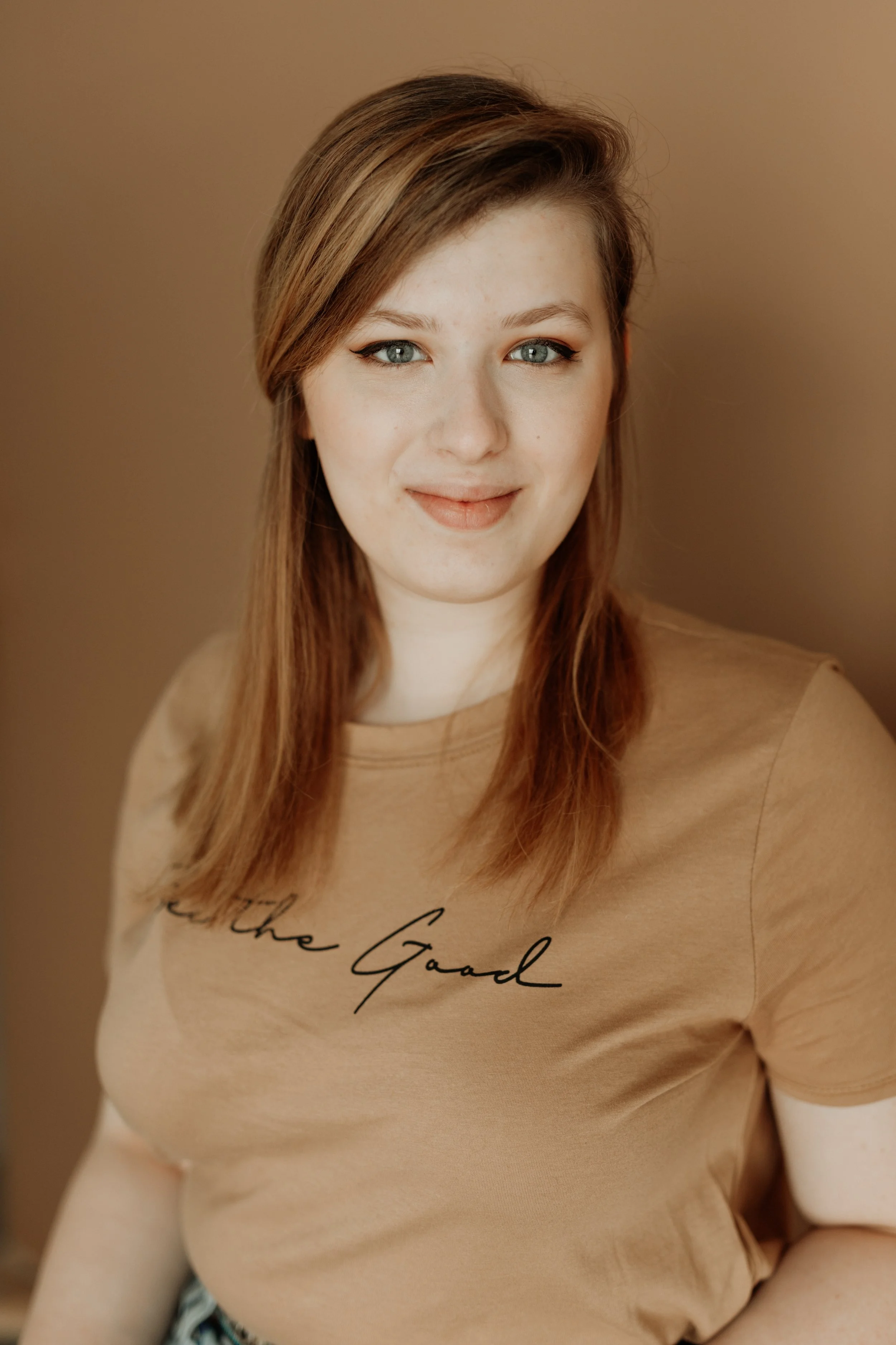 A young woman with light skin, blue eyes, and brown hair with highlights, smiling softly at the camera, wearing a tan T-shirt with black cursive text, against a plain beige background.