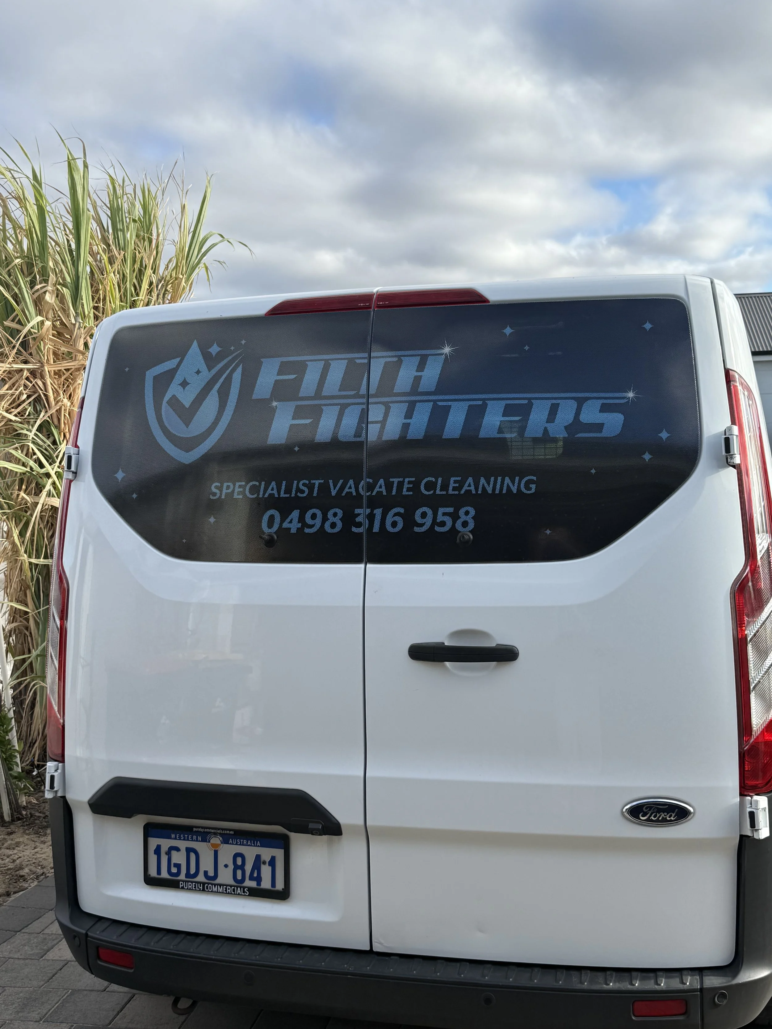 Rear view of a white Ford commercial vehicle with advertising for a cleaning service called 'Filth Fighters' on the back window, including a phone number and the tag 'Specialist Vacate Cleaning,' parked on a pavement near tall grass and a building under a partly cloudy sky.