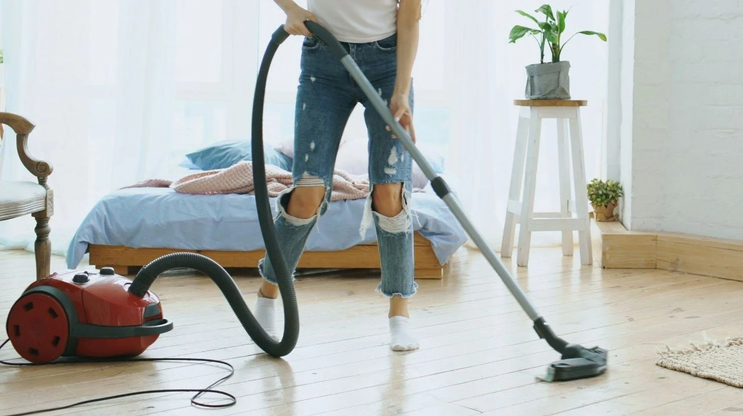Person vacuuming wooden floor in bright bedroom with bed, plants, and white walls.