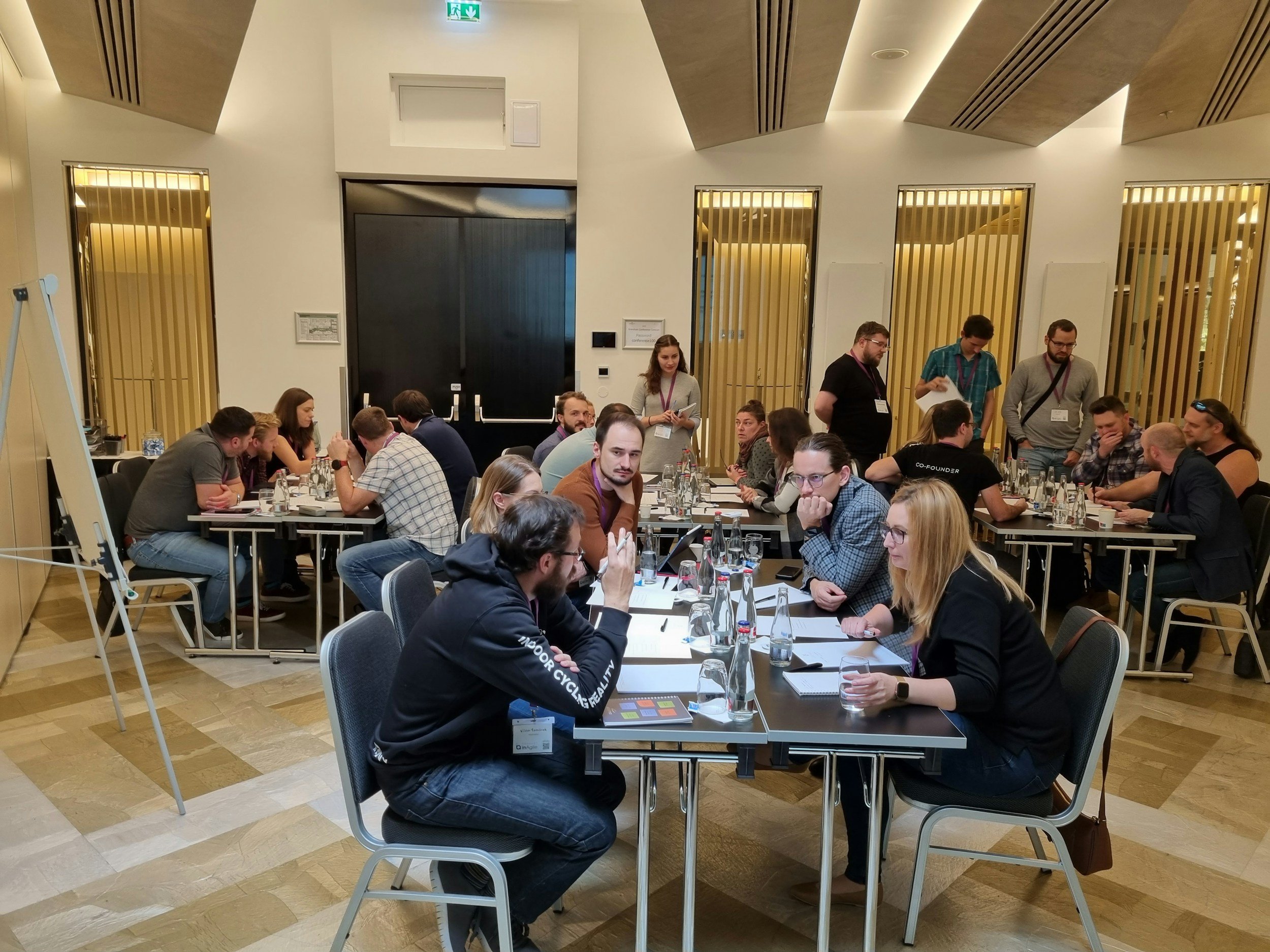 A group of people sitting and standing around conference tables in a modern room, engaged in discussions, with some writing or reading. The room has wooden accent walls and a large, dark elevator door at the front.