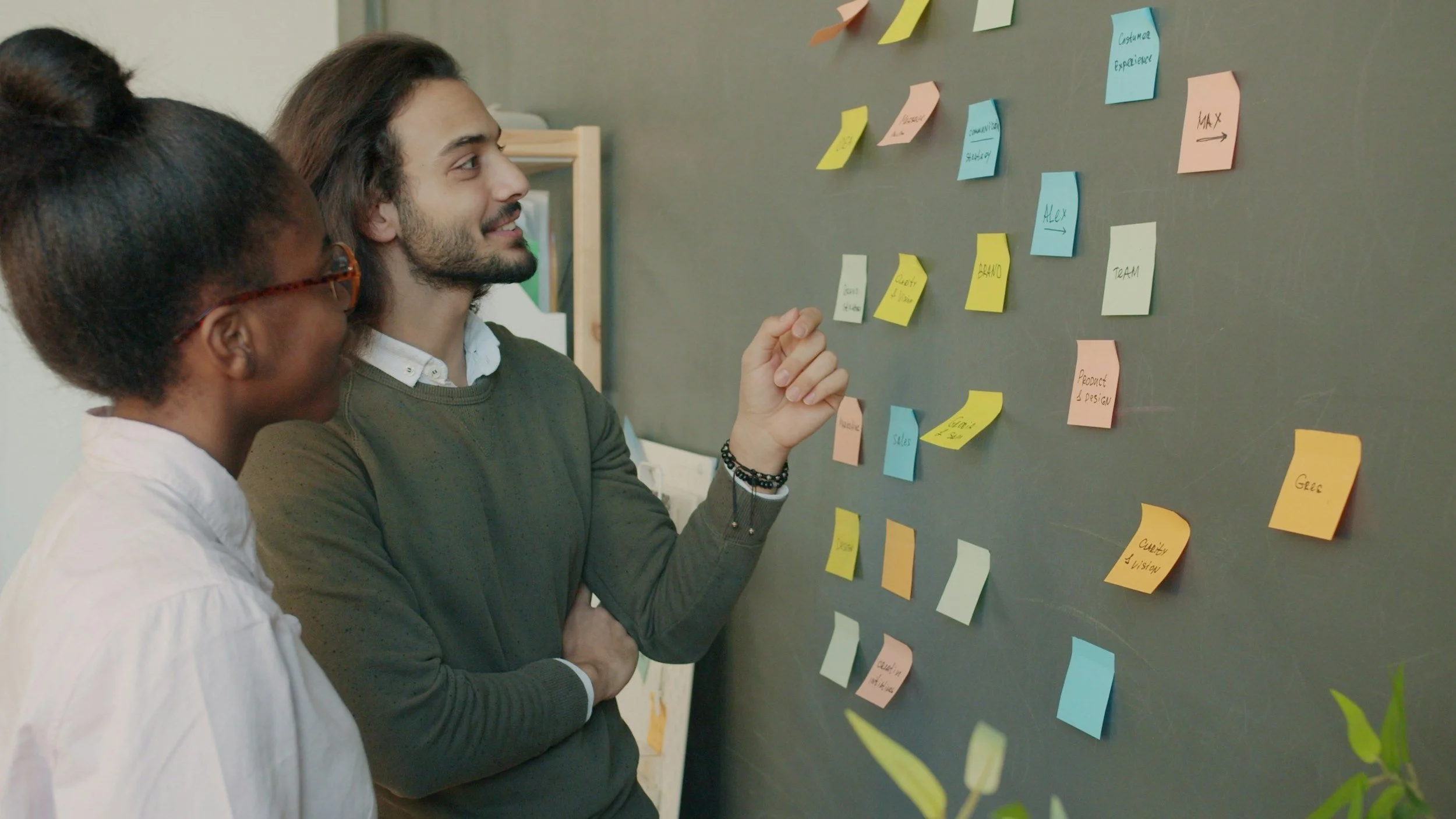 Two people, a woman and a man, analyse colorful sticky notes on a gray wall, likely working together on a project or brainstorming session.