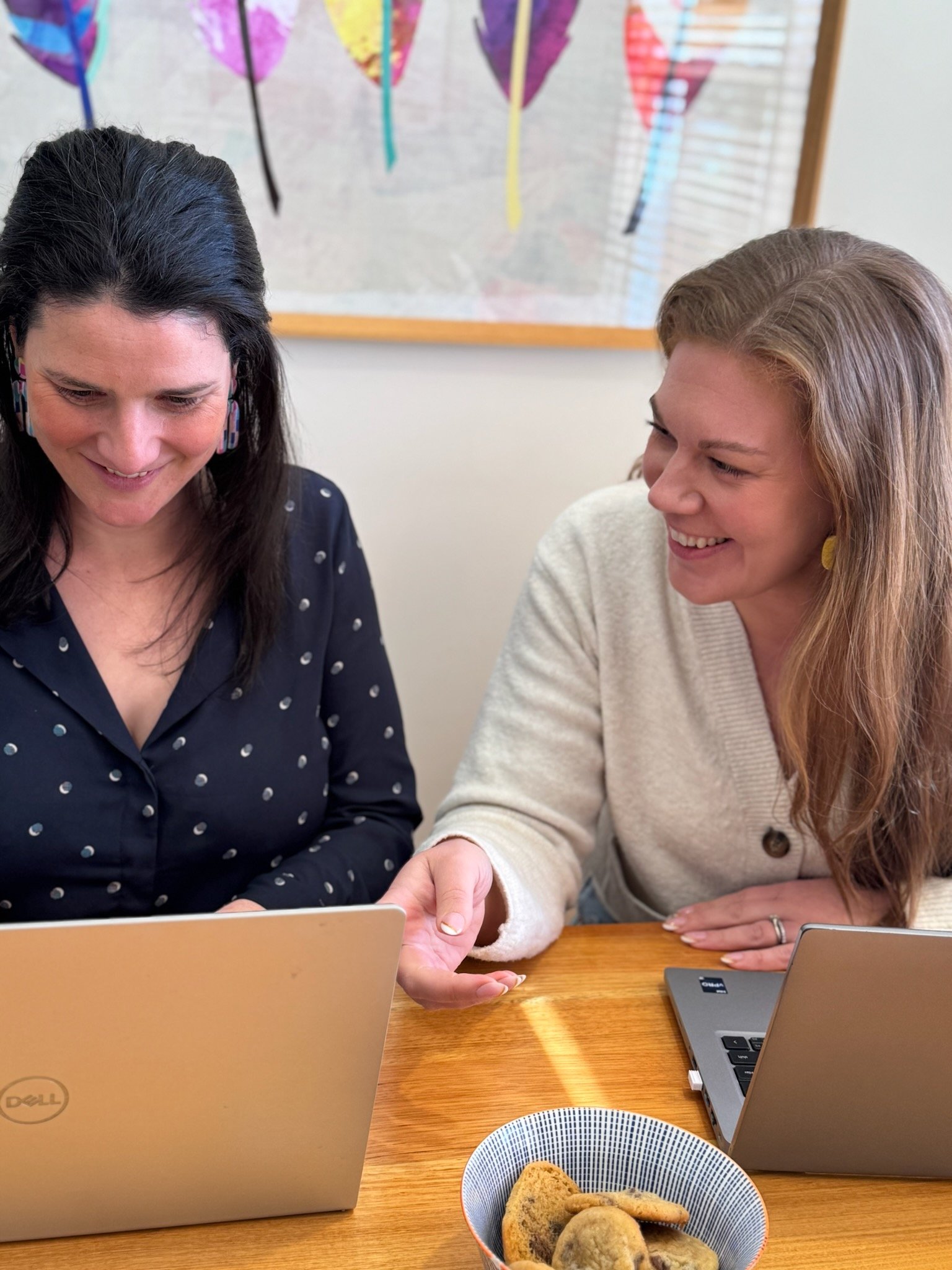 Two women are sitting at a wooden table, looking at a laptop screen and smiling. One woman is wearing a black shirt with white polka dots and has dark hair. The other woman is wearing a light-colored sweater and has blonde hair. On the table, there is a bowl of cookies and two laptops.